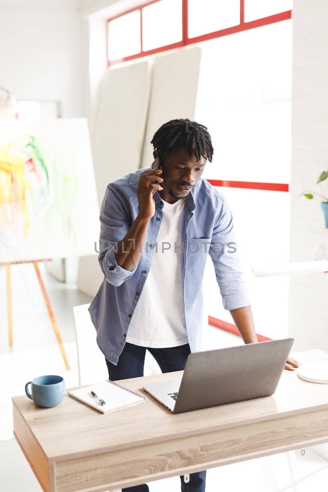 African american male painter at work using laptop and talking on smartphone in art studio. creation and inspiration at an artists painting studio.