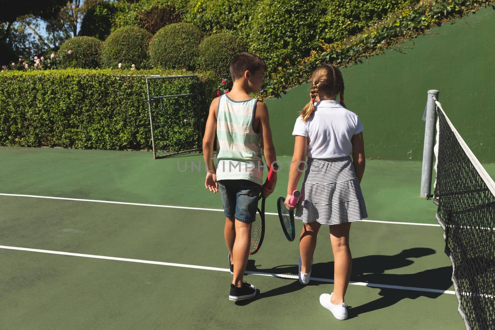 Caucasian boy and girl outdoors, holding tennis rackets and walking on tennis court by Wavebreakmedia