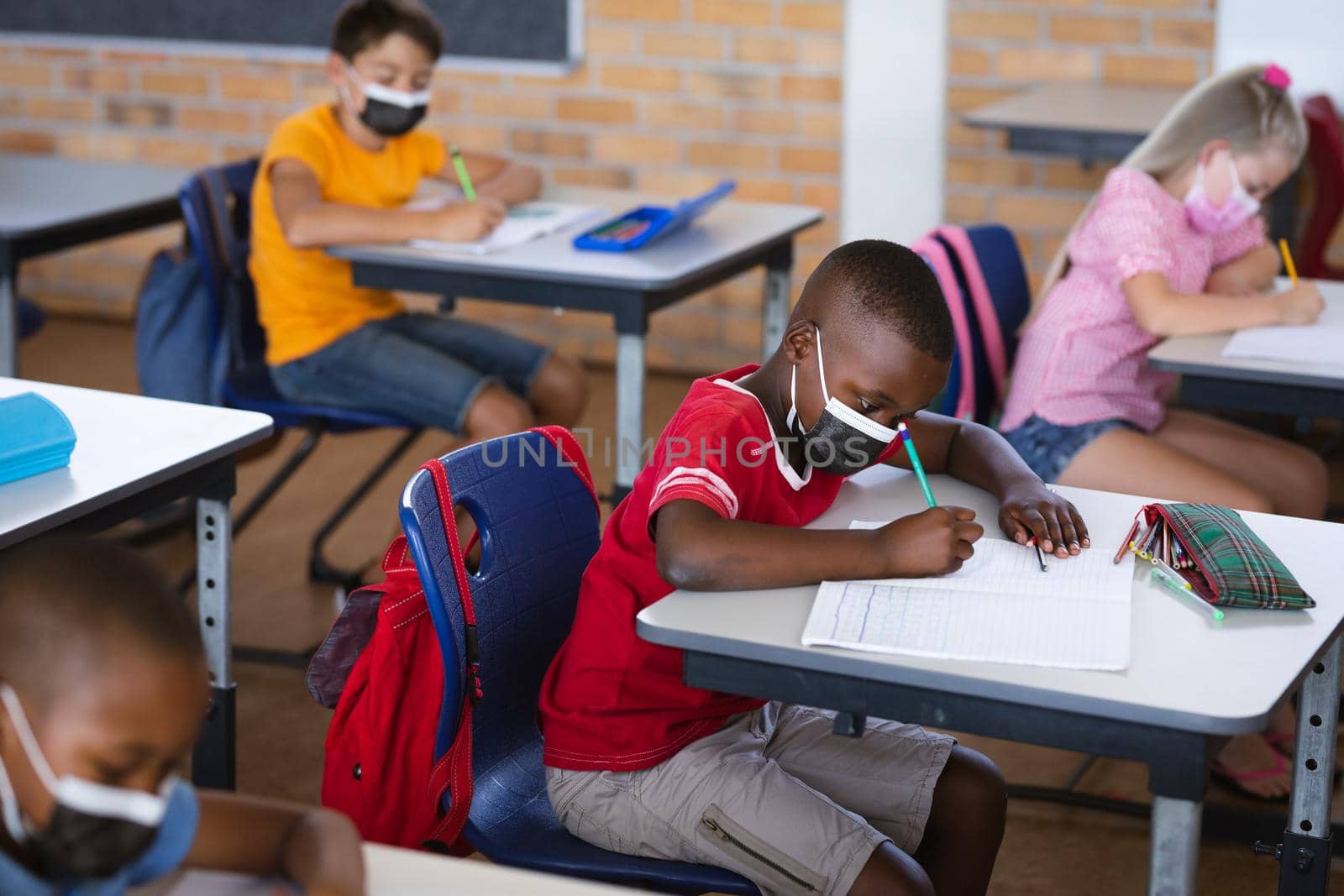 African american boy wearing face mask studying while sitting on his desk in the class at school by Wavebreakmedia