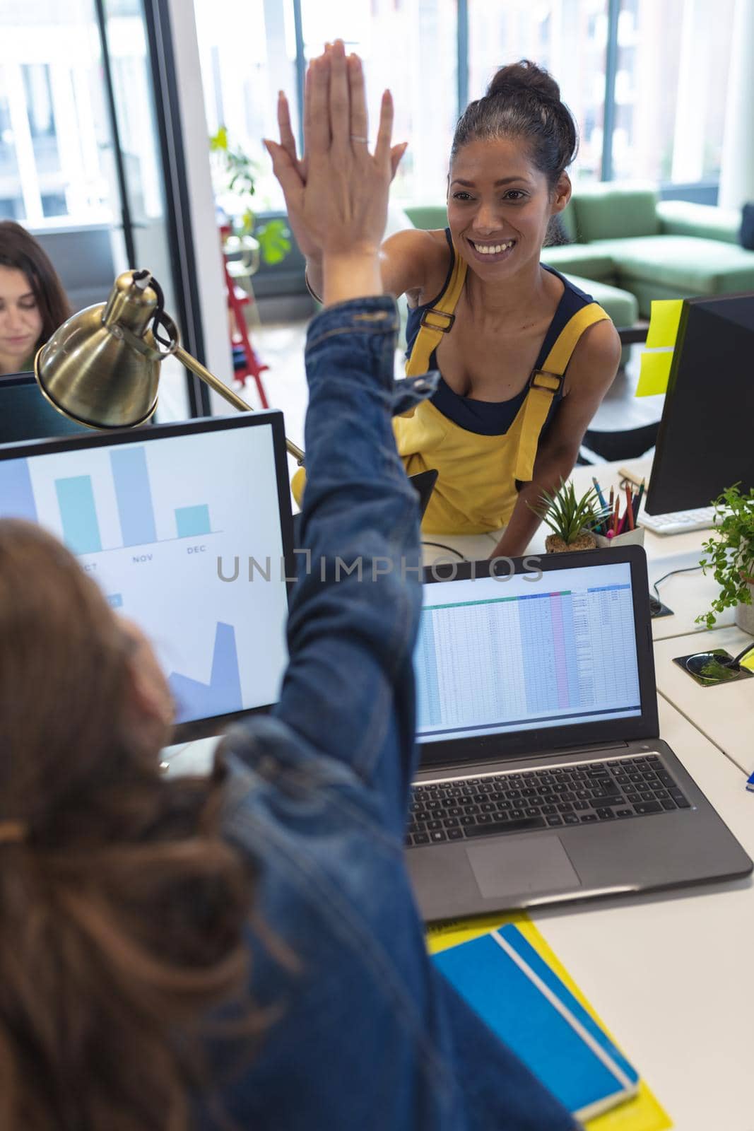 Two diverse female creative colleagues sitting at desk high-fiving and smiling. modern office of a creative design business.
