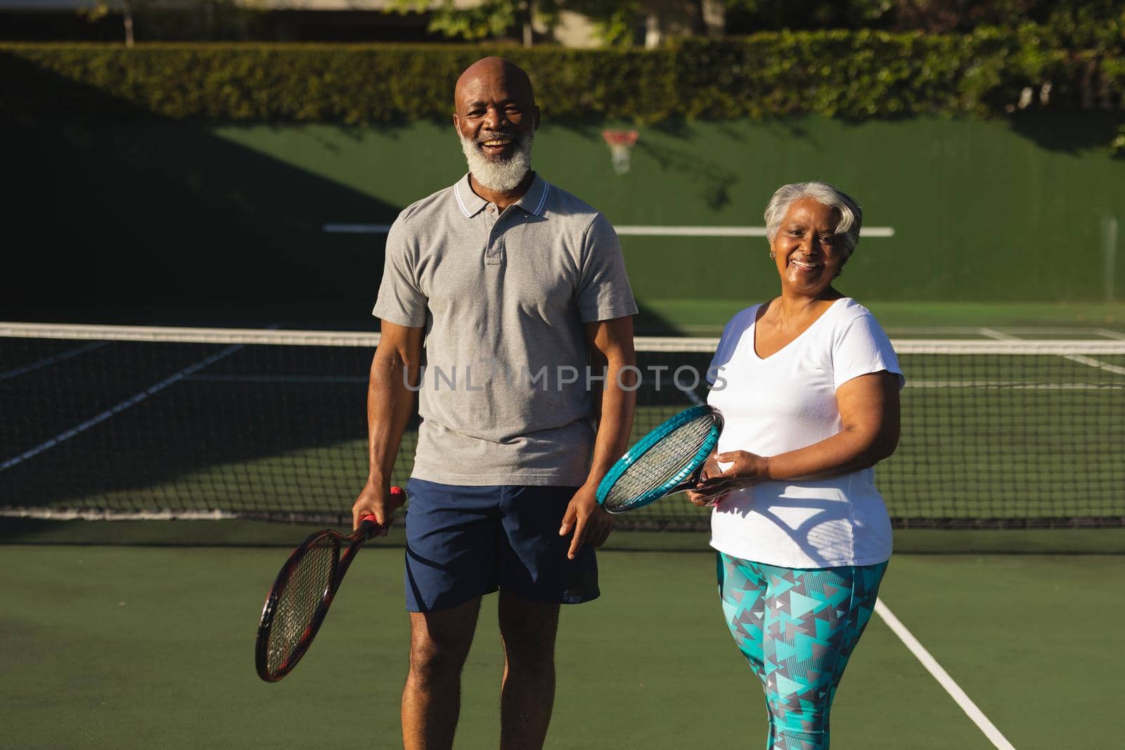 Portrait of smiling senior african american couple with tennis rackets on tennis court. retirement and active senior lifestyle concept.