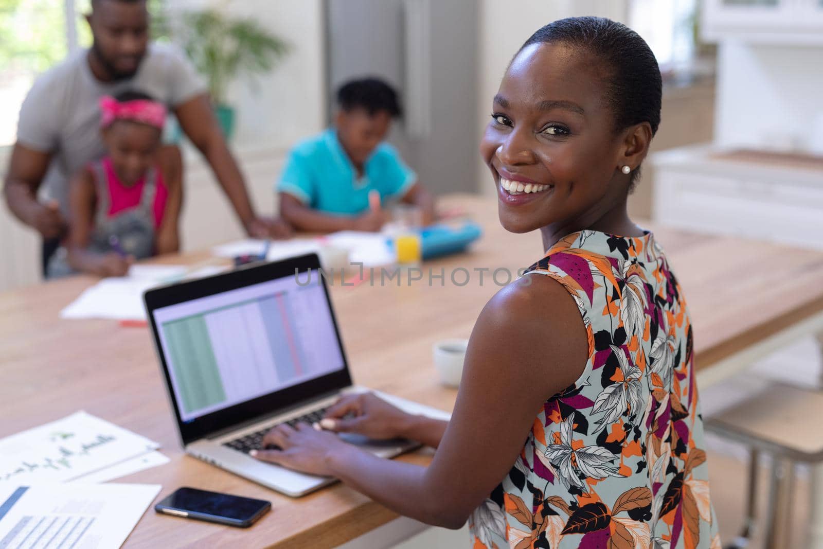 Portrait of smiling african american mother working at home using laptop with family in background. staying at home in isolation during quarantine lockdown.