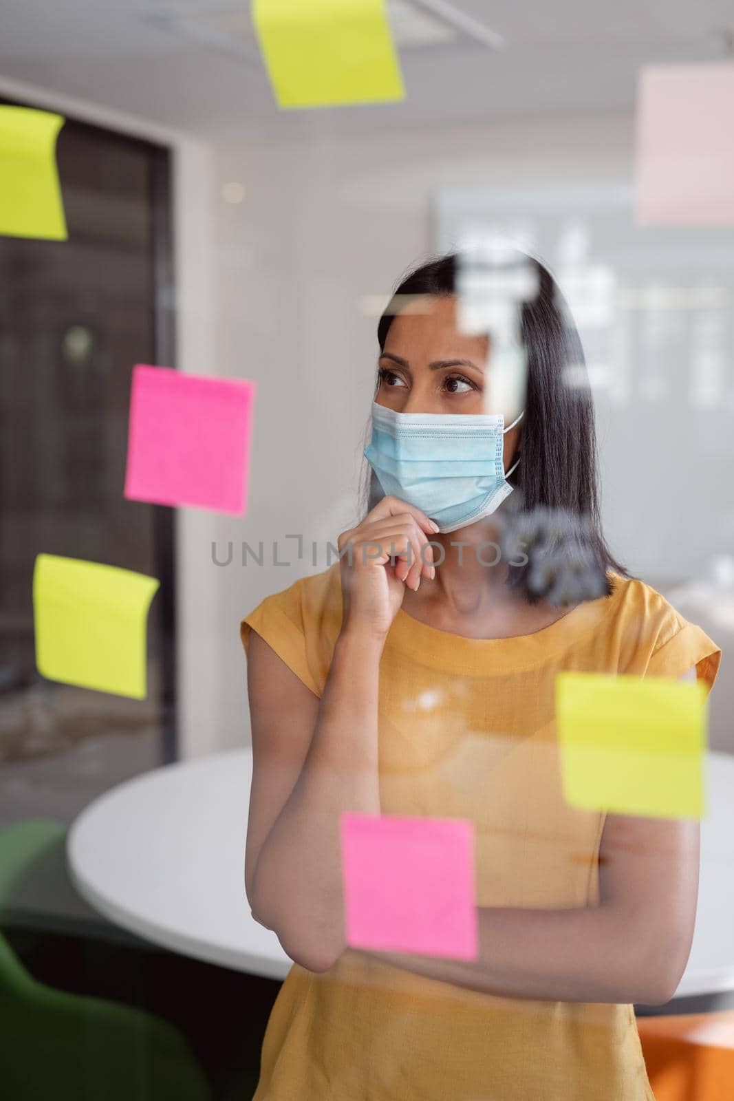 Caucasian businesswoman wearing face mask brainstorming, reading colourful memo notes on glass wall. working in business at a modern office during coronavirus covid 19 pandemic.