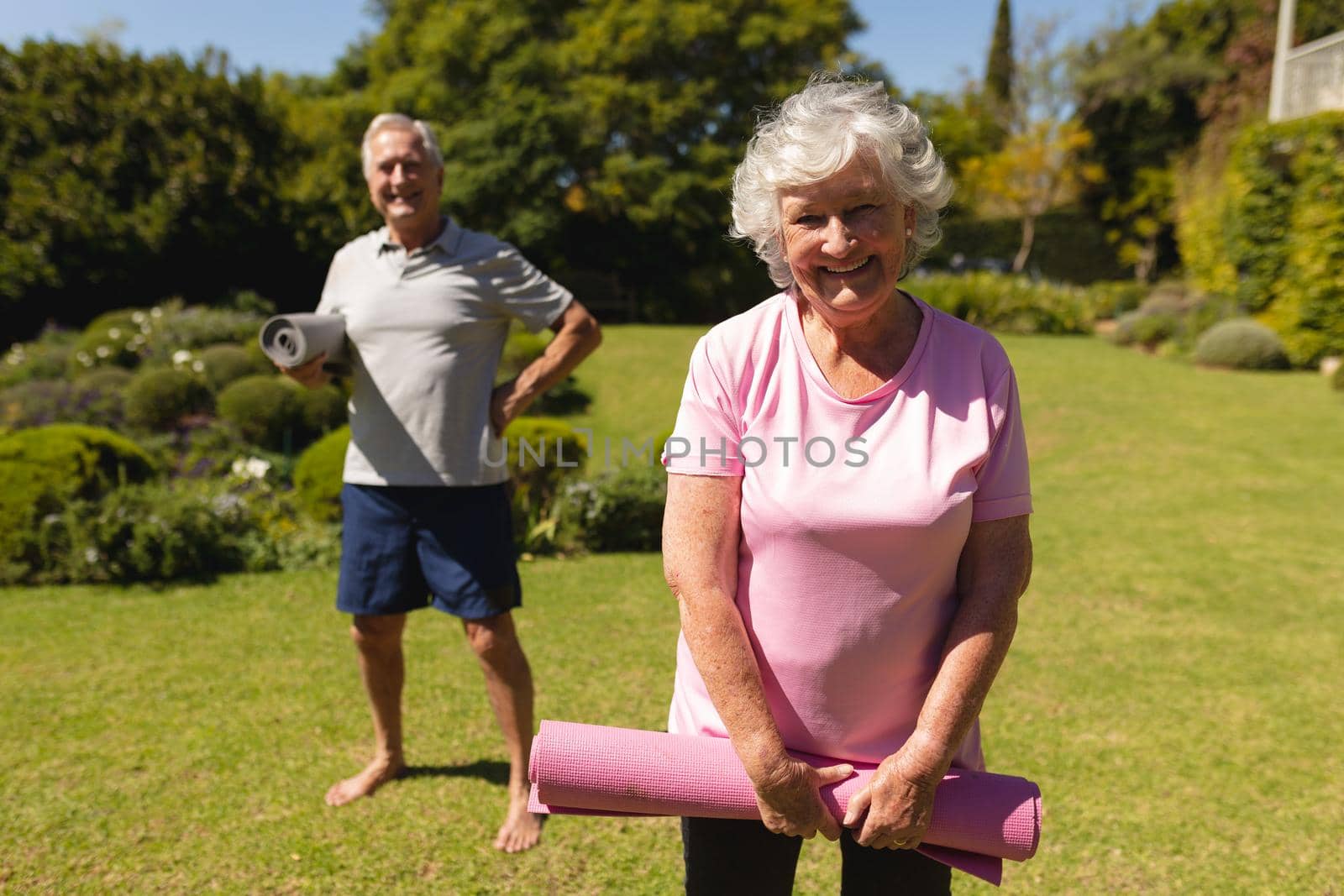 Portrait of senior caucasian couple holding yoga mats, looking at camera and smiling in sunny garden. retirement retreat and active senior lifestyle concept.