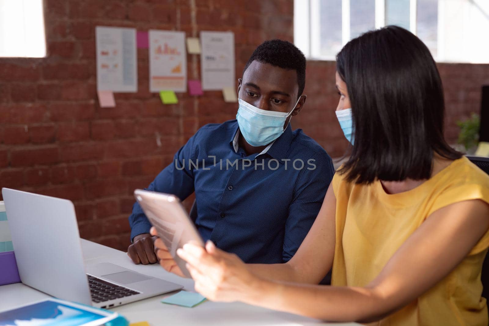 Diverse male and female colleague in face masks sitting at desk discussing, using laptop and tablet. working in business at a modern office during coronavirus covid 19 pandemic.