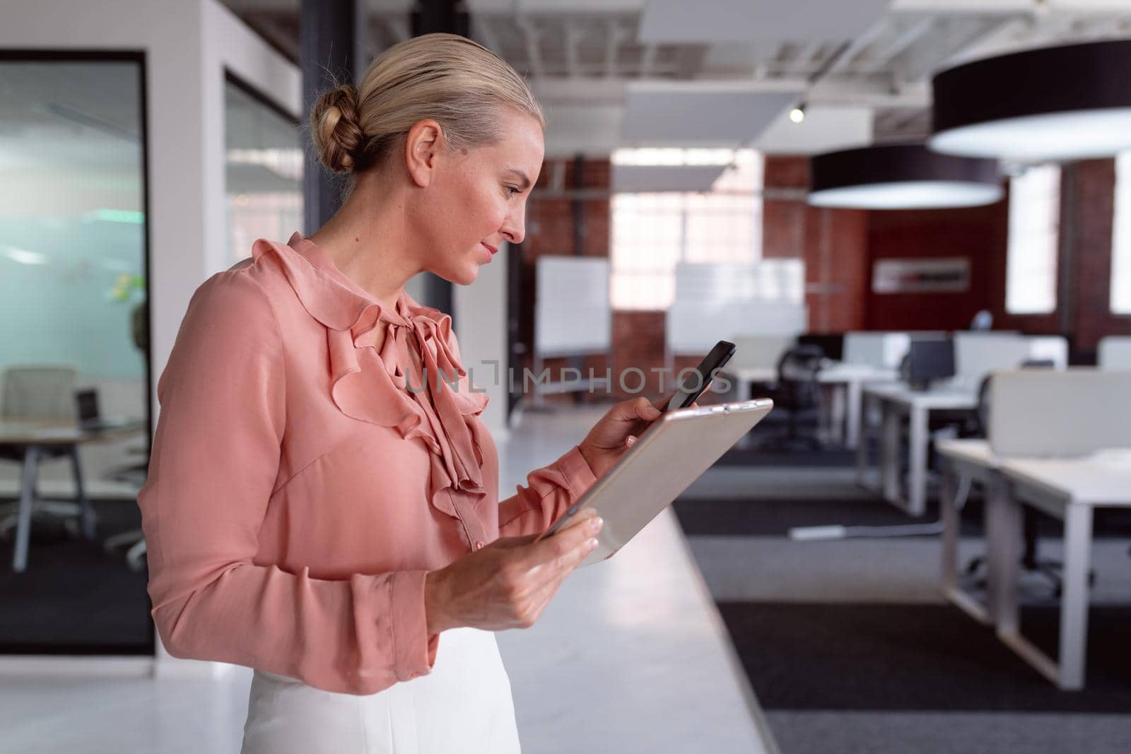 Happy caucasian businesswoman standing in office holding tablet and using smartphone by Wavebreakmedia