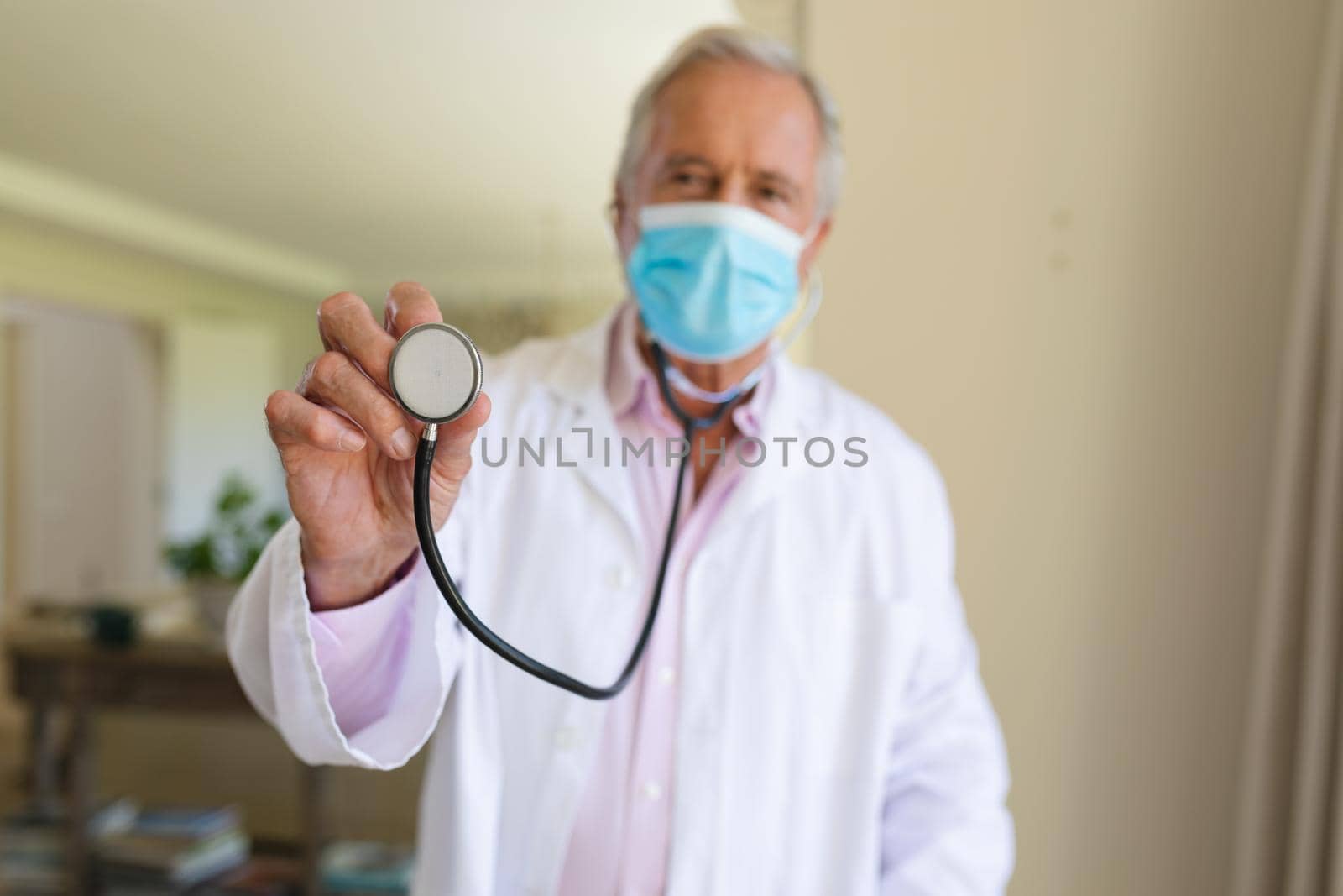 Portrait of senior caucasian male doctor wearing face mask holding stethoscope looking at camera. medicine and healthcare services during covid 19 pandemic concept.