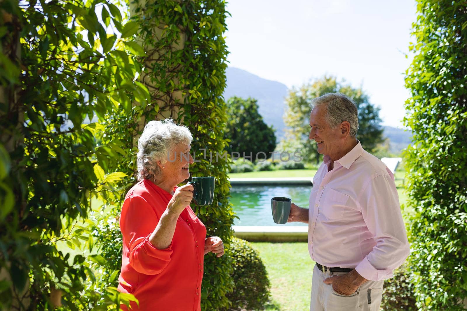 Senior caucasian couple smiling and holding mugs in sunny garden. retreat, retirement and happy senior lifestyle concept.