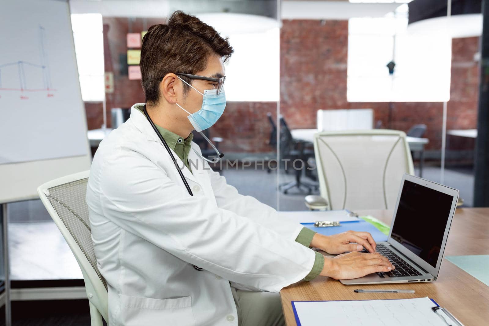 Asian male doctor wearing face mask sitting at desk in hospital office using laptop. medical and healthcare services during coronavirus covid 19 pandemic.