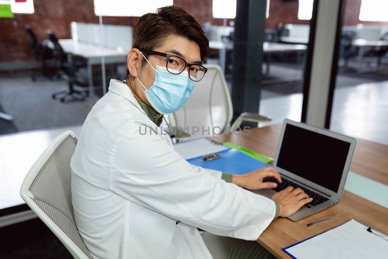 Asian male doctor wearing face mask sitting at desk in hospital office using laptop by Wavebreakmedia