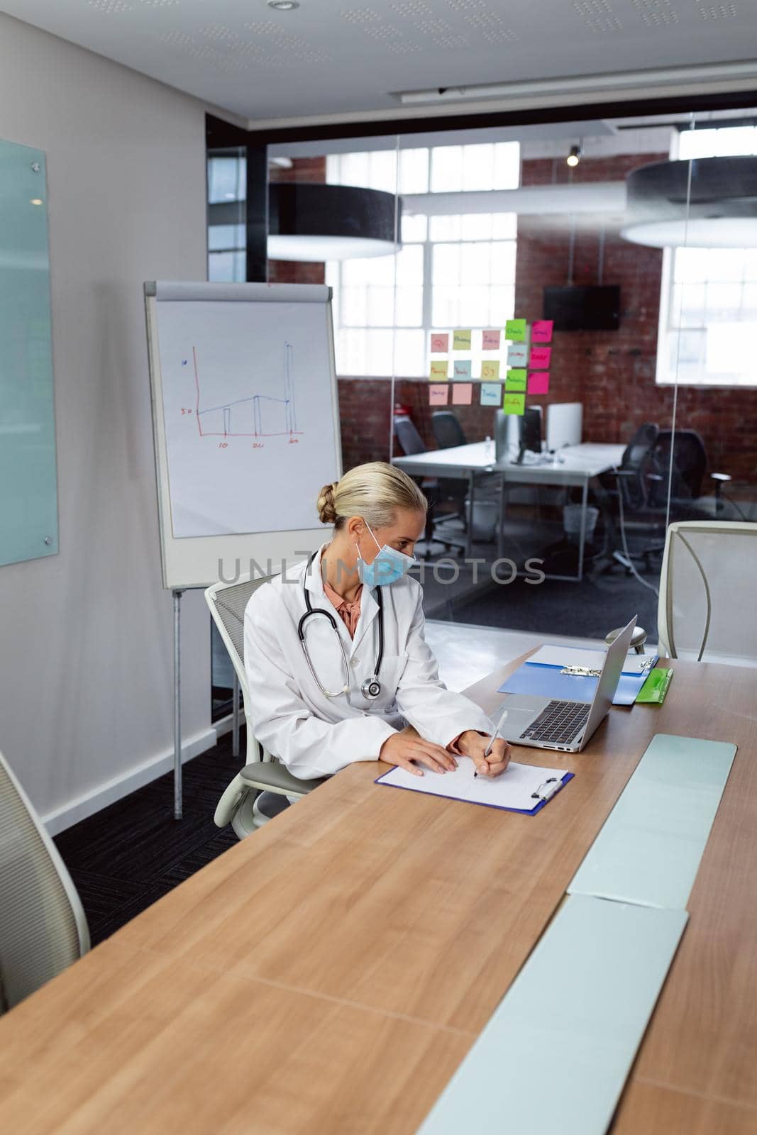 Caucasian female doctor wearing face mask sitting in hospital office using laptop doing paperwork by Wavebreakmedia