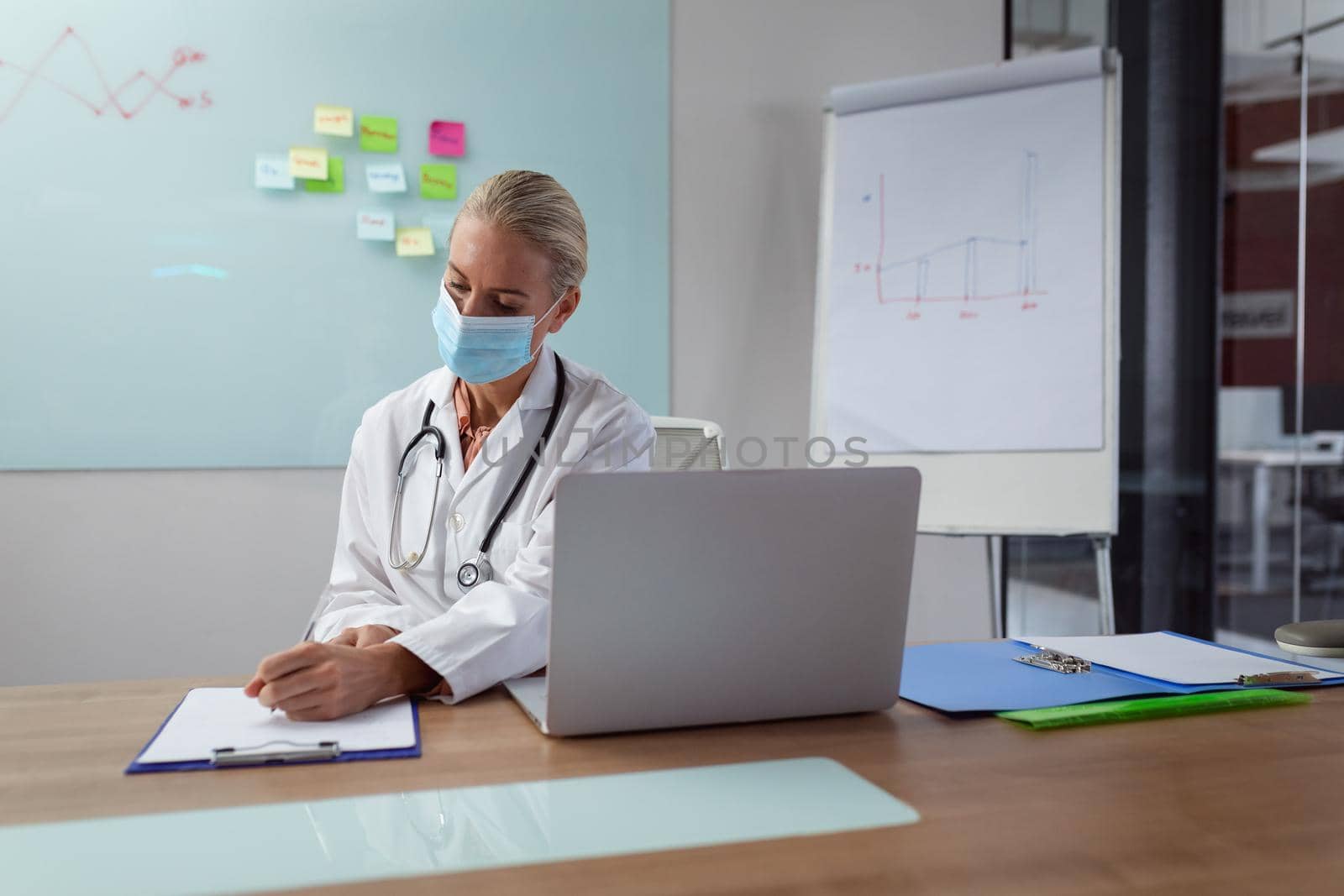 Caucasian female doctor wearing face mask sitting at desk working in hospital office by Wavebreakmedia