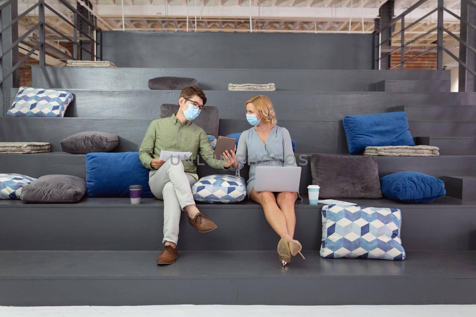 Male and female colleague in face masks sitting in foyer discussing and using tablet and laptop. working in business at a modern office during coronavirus covid 19 pandemic.