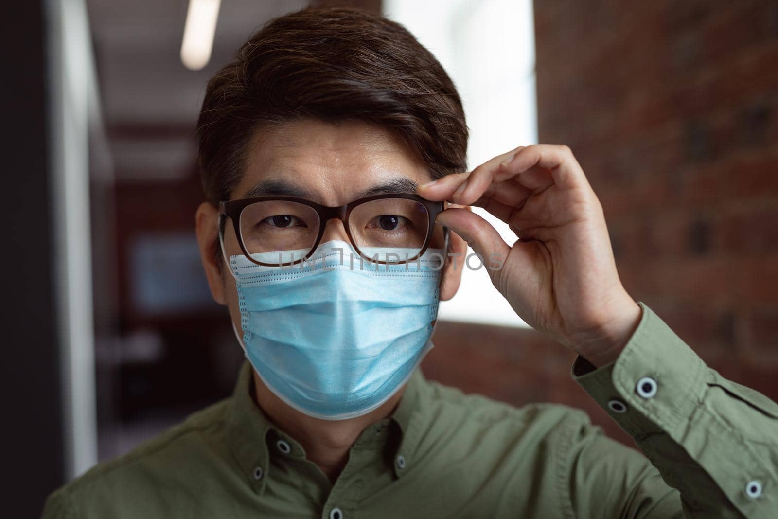 Portrait of asian businessman wearing face mask in office holding glasses and looking to camera by Wavebreakmedia