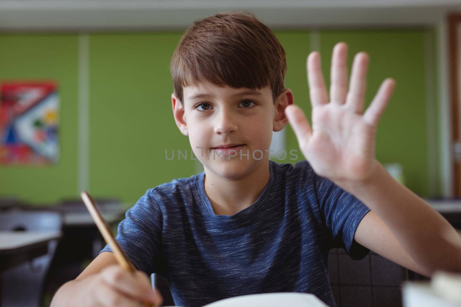 Portrait of caucasian schoolboy in classroom sitting at desk and waving. childhood and education at elementary school.