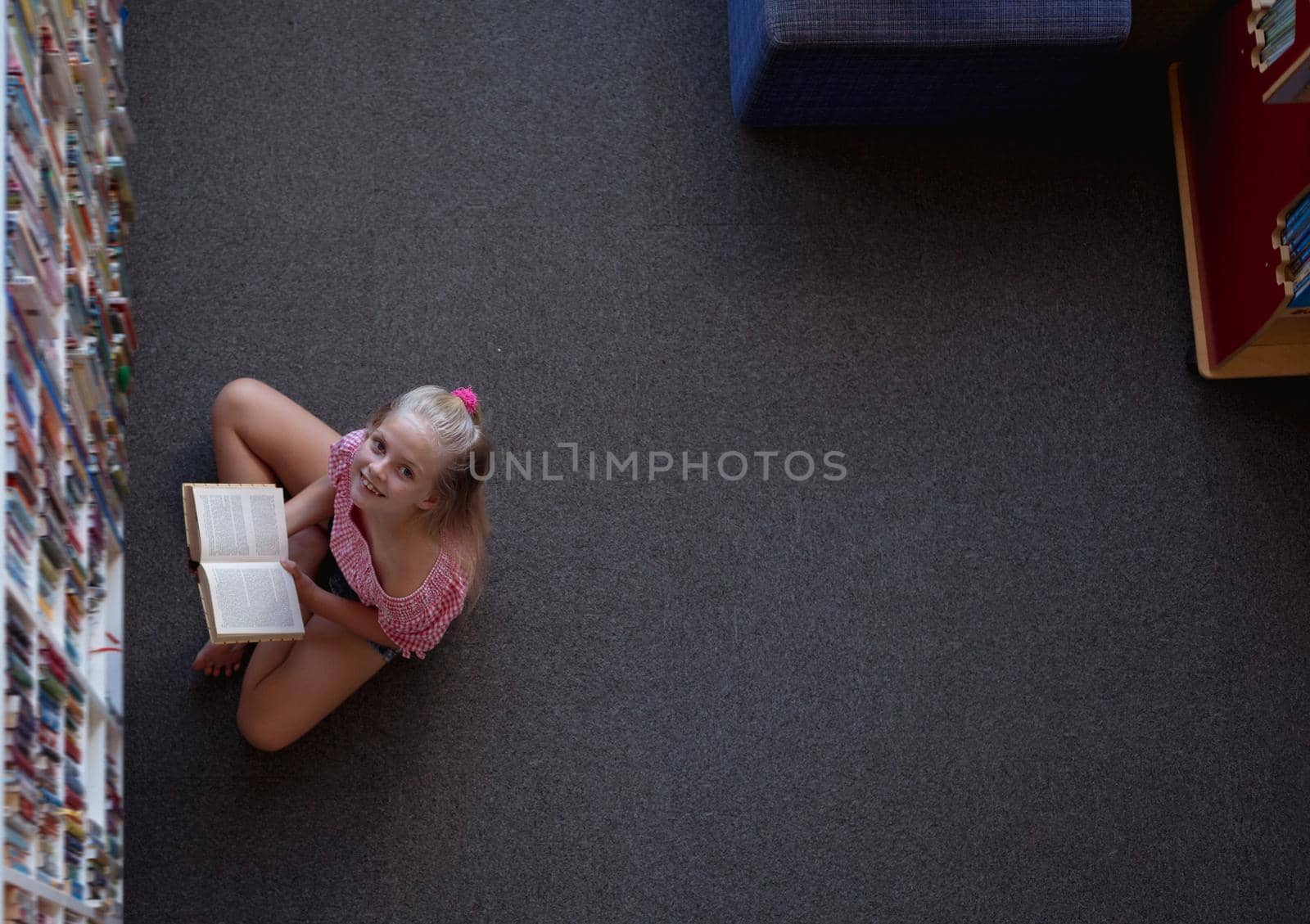 Overhead view of smiling caucasian schoolgirl sitting on floor reading book in school library. childhood and education at elementary school.