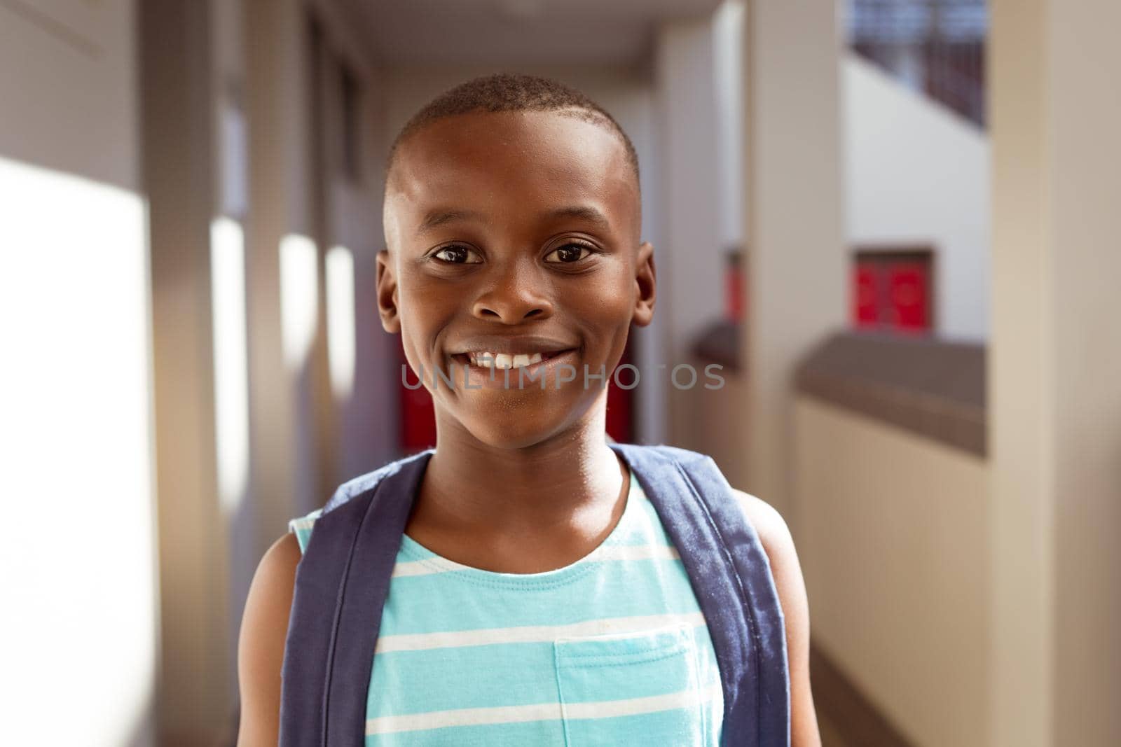 Portrait of smiling african american schoolboy wearing schoolbag standing in school corridor by Wavebreakmedia