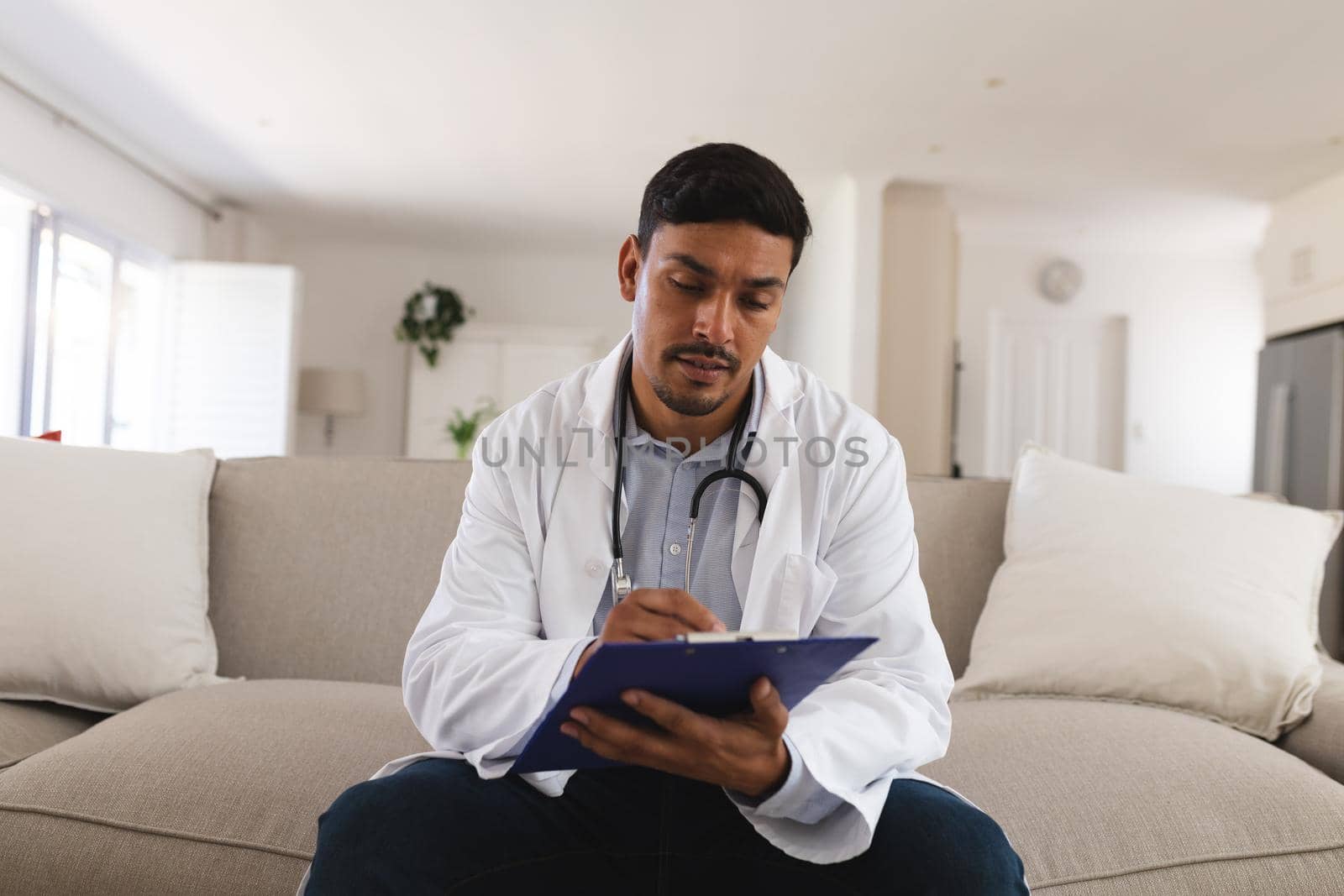 Hispanic male doctor sitting on couch making notes during consultation video call by Wavebreakmedia