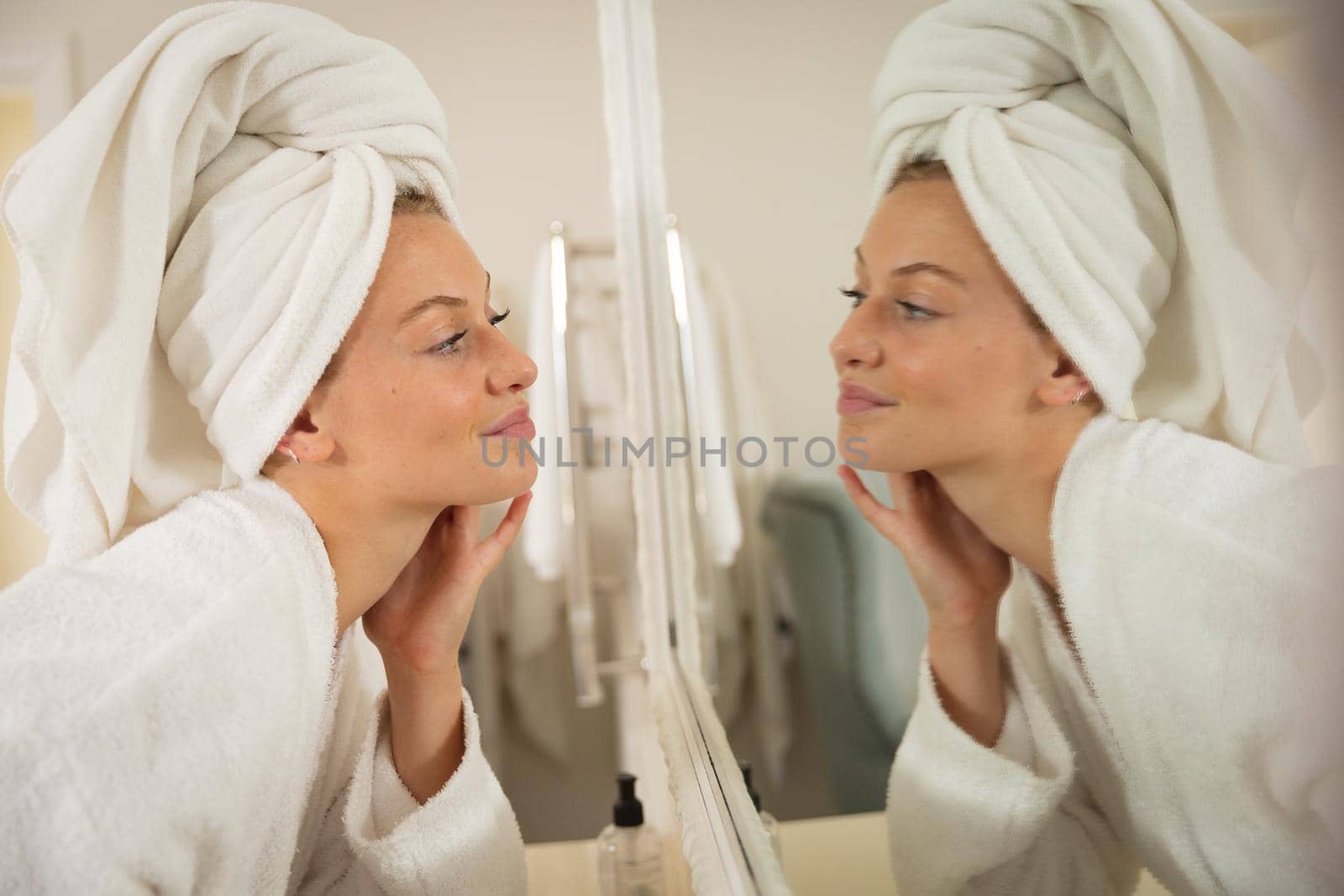 Smiling caucasian woman in bathroom with towel on head, looking in mirror and moisturising face by Wavebreakmedia
