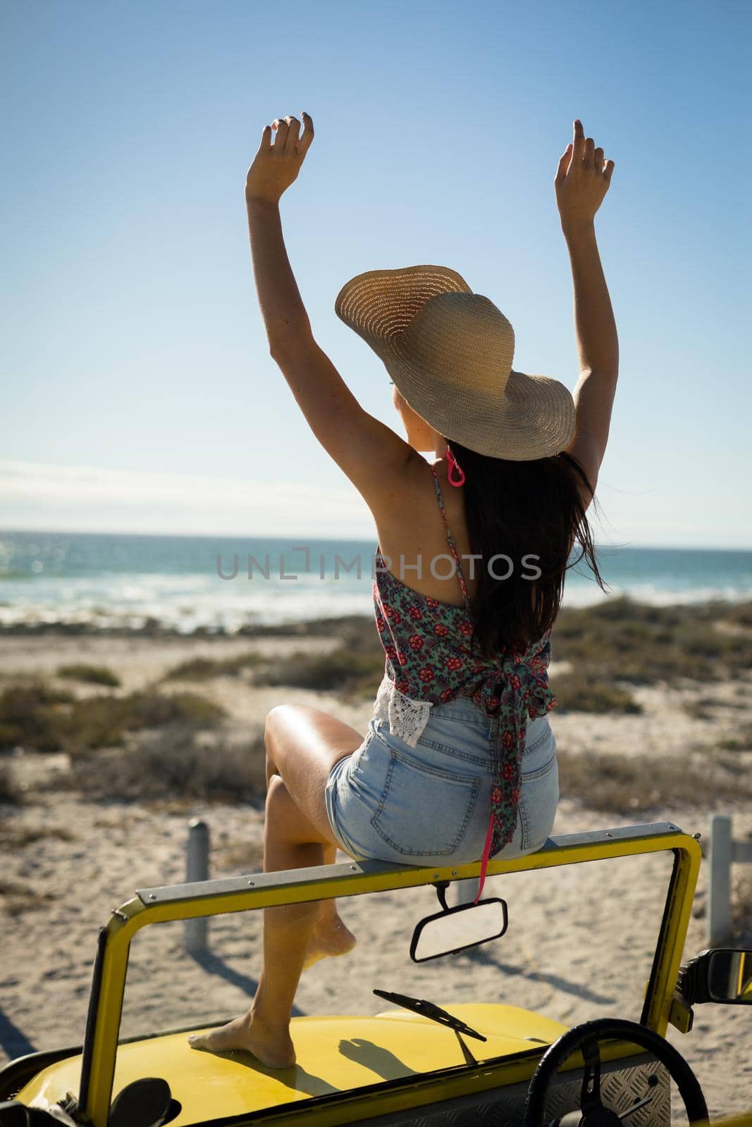 Caucasian woman sitting on beach buggy by the sea wearing straw hat looking toward sea with hands up. beach break on summer holiday road trip.