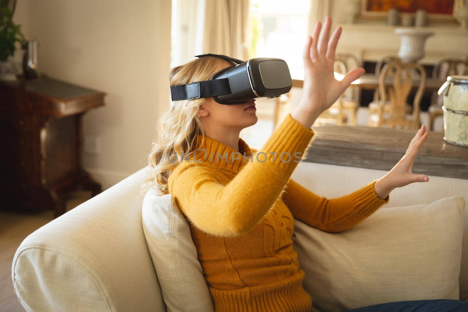 Caucasian woman sitting on couch in luxury living room wearing vr headset, with hands raised. spending free time at home with technology.