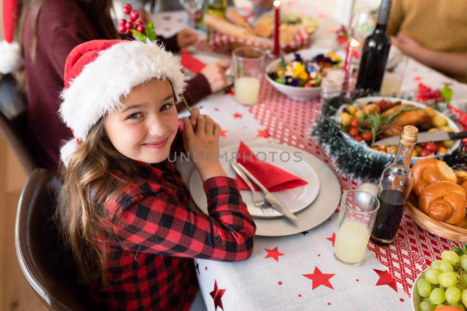 Happy caucasian girl wearing santa hat sitting at christmas table and looking at camera. family christmas time and festivity together at home.