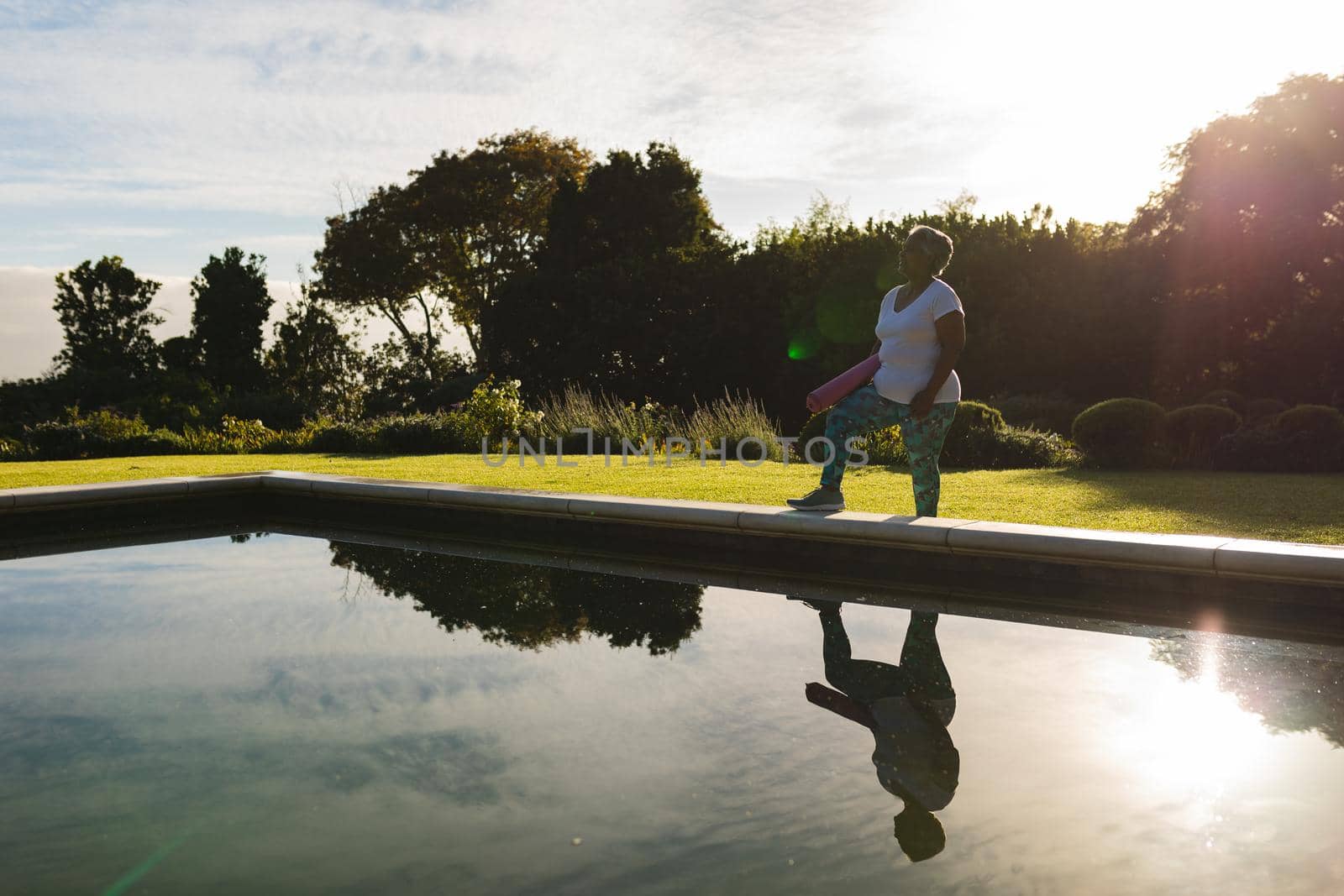 Senior african american woman standing with yoga mat by outdoor pool in countryside. retirement and active senior lifestyle concept.