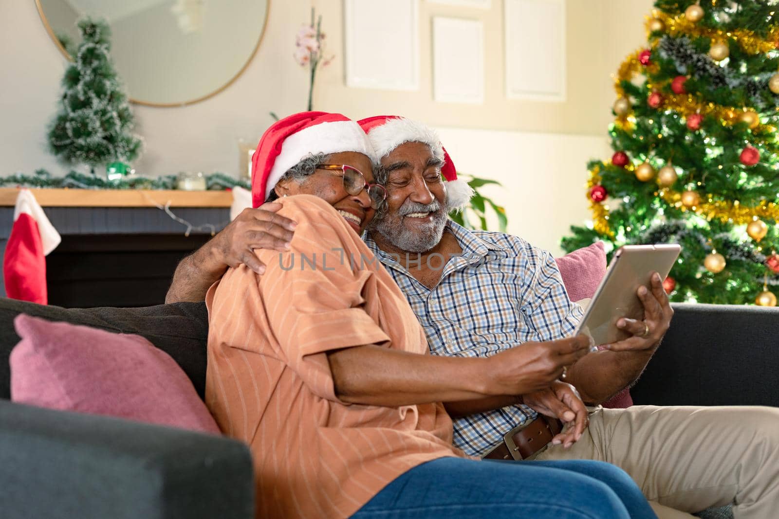 Happy african american couple wearing santa hats using tablet, having video call at christmas time. christmas, festivity and communication technology at home.
