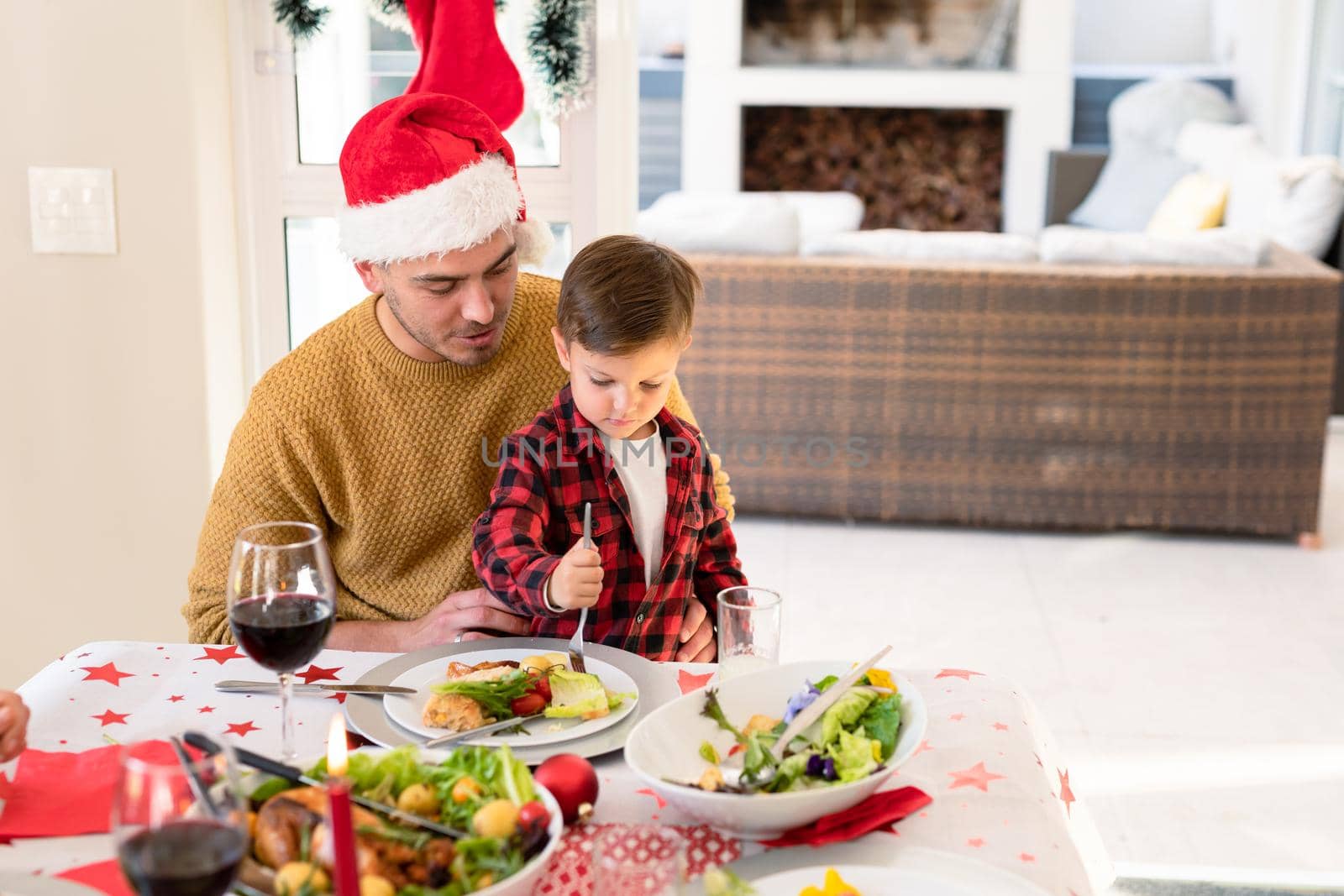 Happy caucasian father and son wearing santa hats sitting at christmas table. family christmas time and festivity together at home.