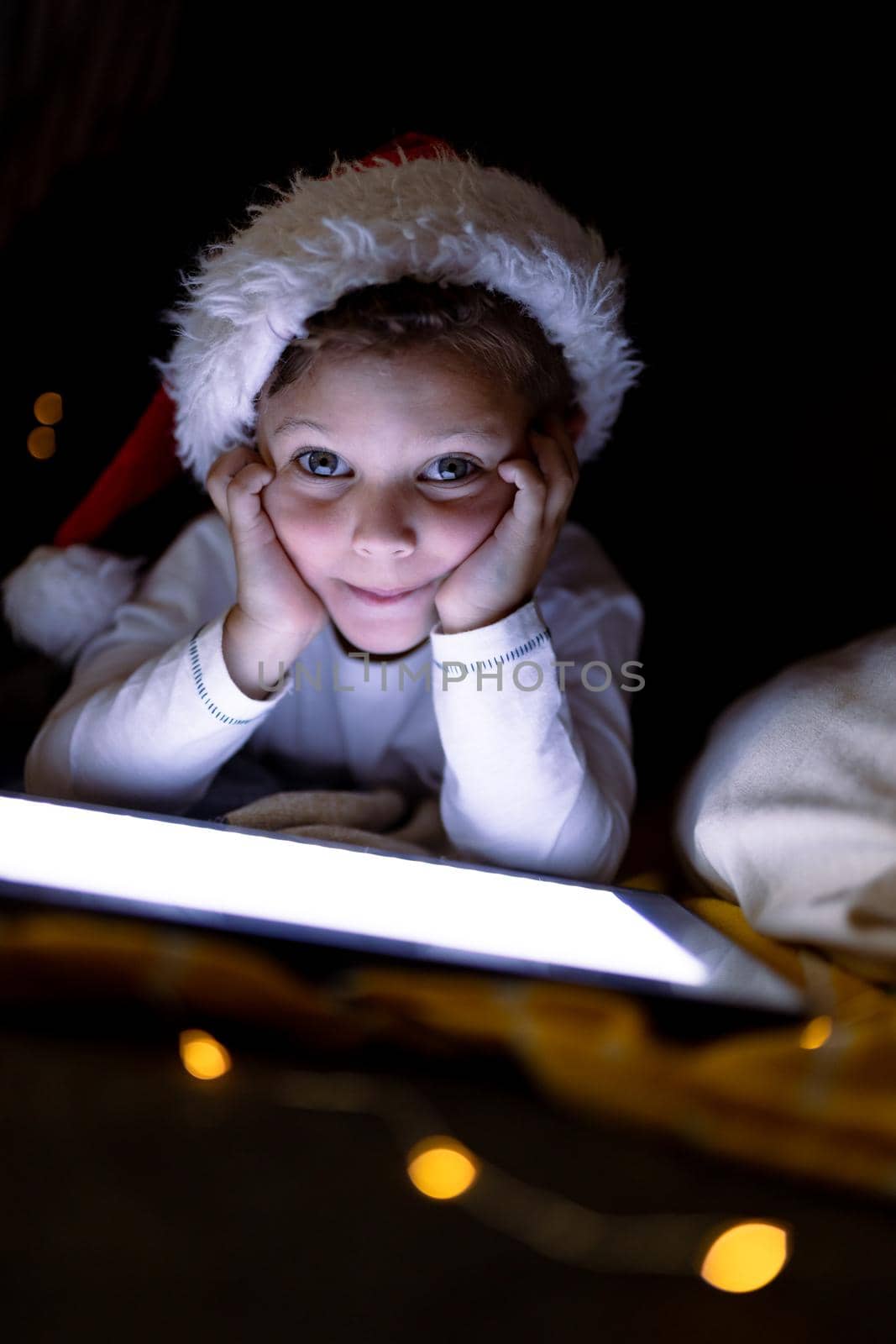 Caucasian boy wearing santa hat, using tablet, looking at camera at christmas time. childhood, chiristmas, festivity and tradition at home.