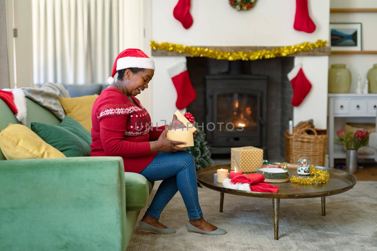Happy african american senior woman in santa hat opening present at christmas time. retirement lifestyle and christmas festivities, celebrating at home.