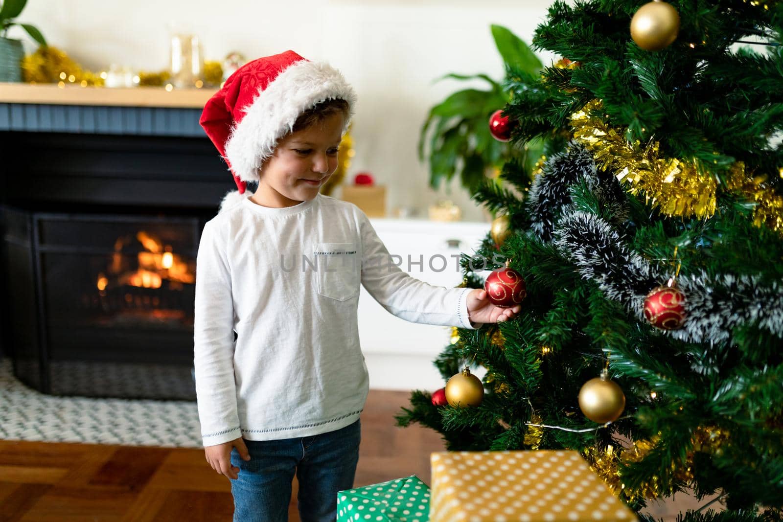 Happy caucasian boy wearing santa hat, decorating christmas tree at christmas time. childhood, chiristmas, festivity and tradition at home.