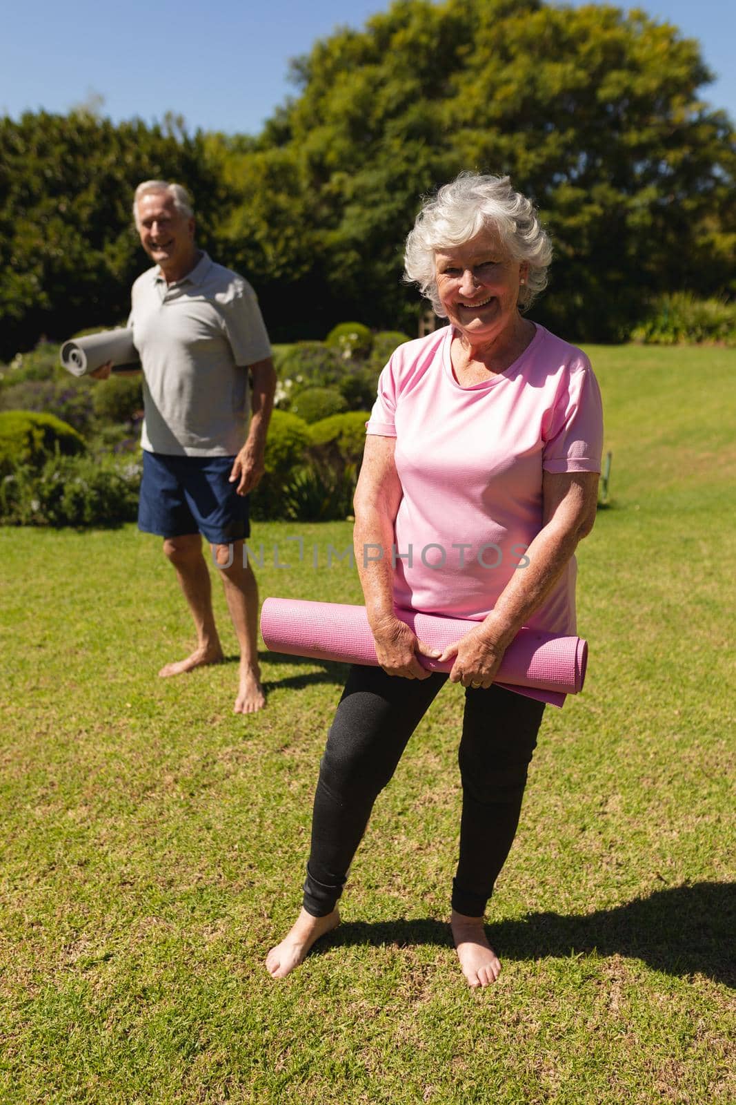 Portrait of senior caucasian couple holding yoga mats, looking at camera and smiling in sunny garden. retirement retreat and active senior lifestyle concept.