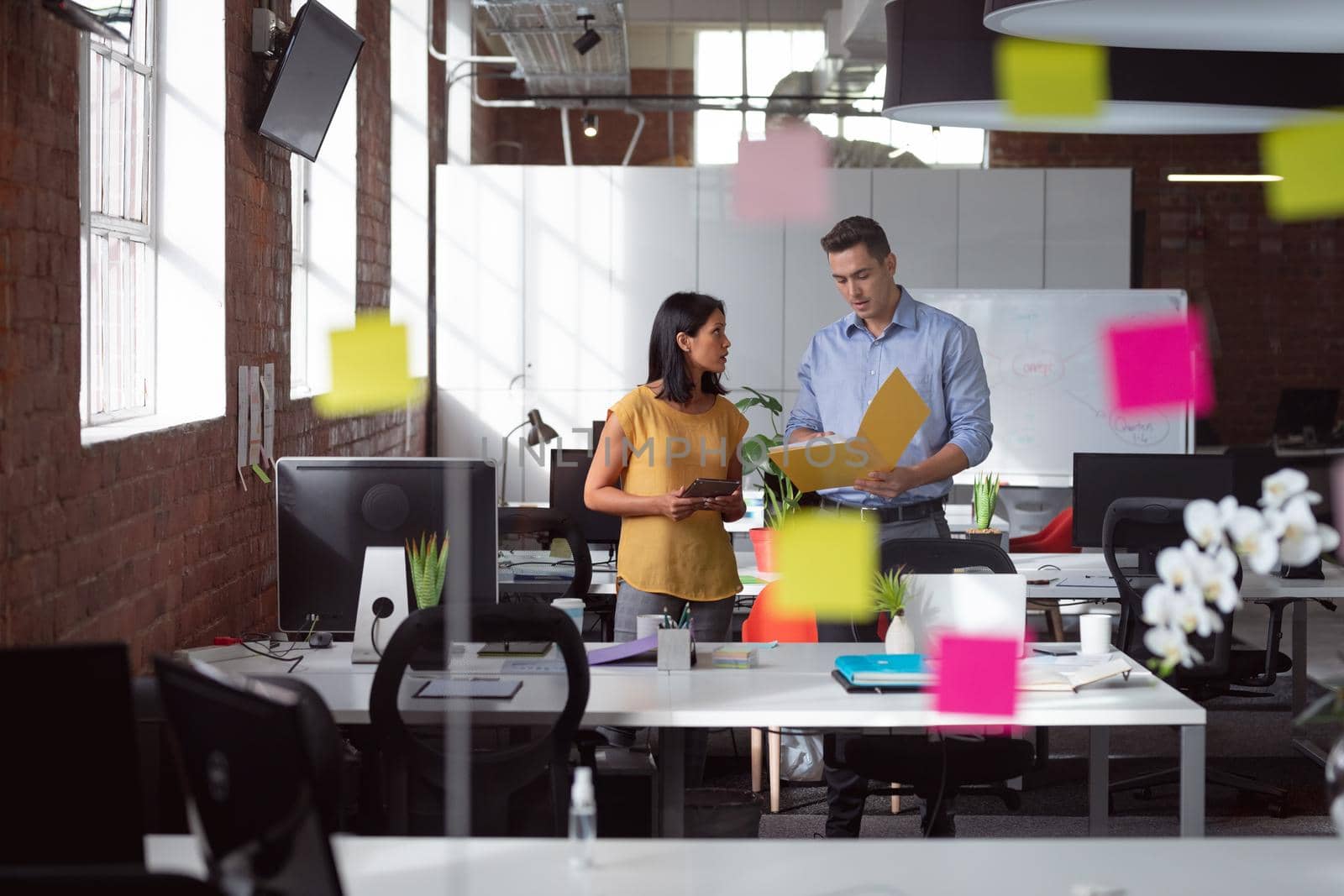 Caucasian male and female colleague discuss tablet and file, view through glass wall and memo notes by Wavebreakmedia