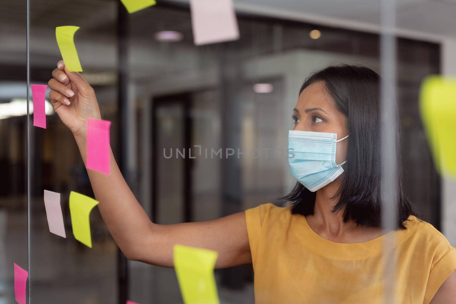 Caucasian businesswoman wearing face mask brainstorming, reading colourful memo notes on glass wall by Wavebreakmedia