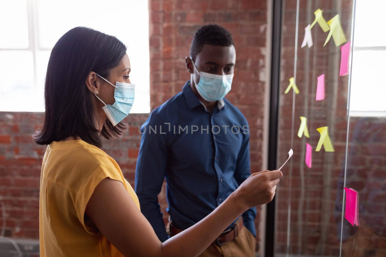 Diverse male and female colleague in face masks brainstorming, reading memo notes on glass wall. working in business at a modern office during coronavirus covid 19 pandemic.