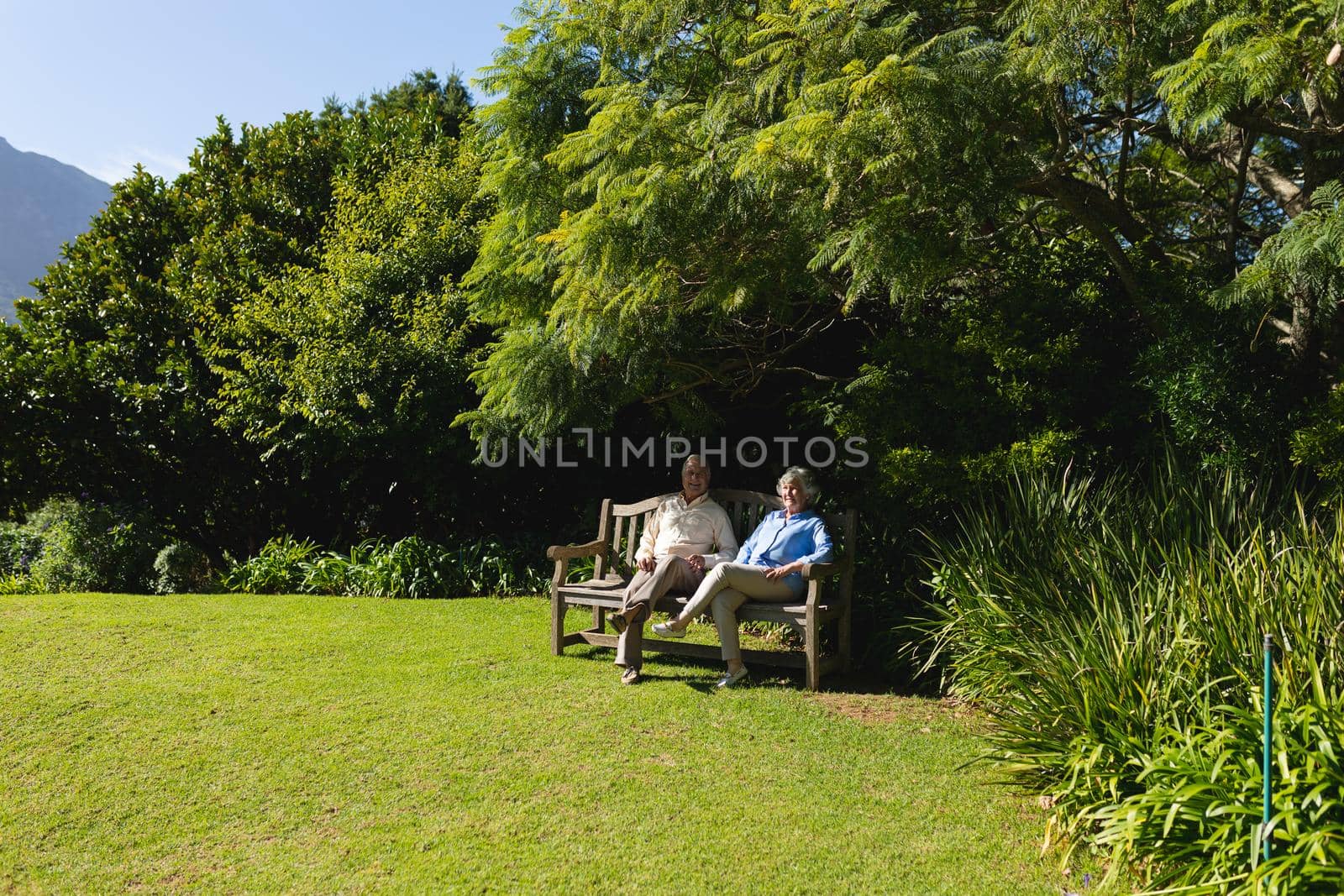 Senior caucasian couple sitting on bench together in sunny garden. retreat, retirement and happy senior lifestyle concept.