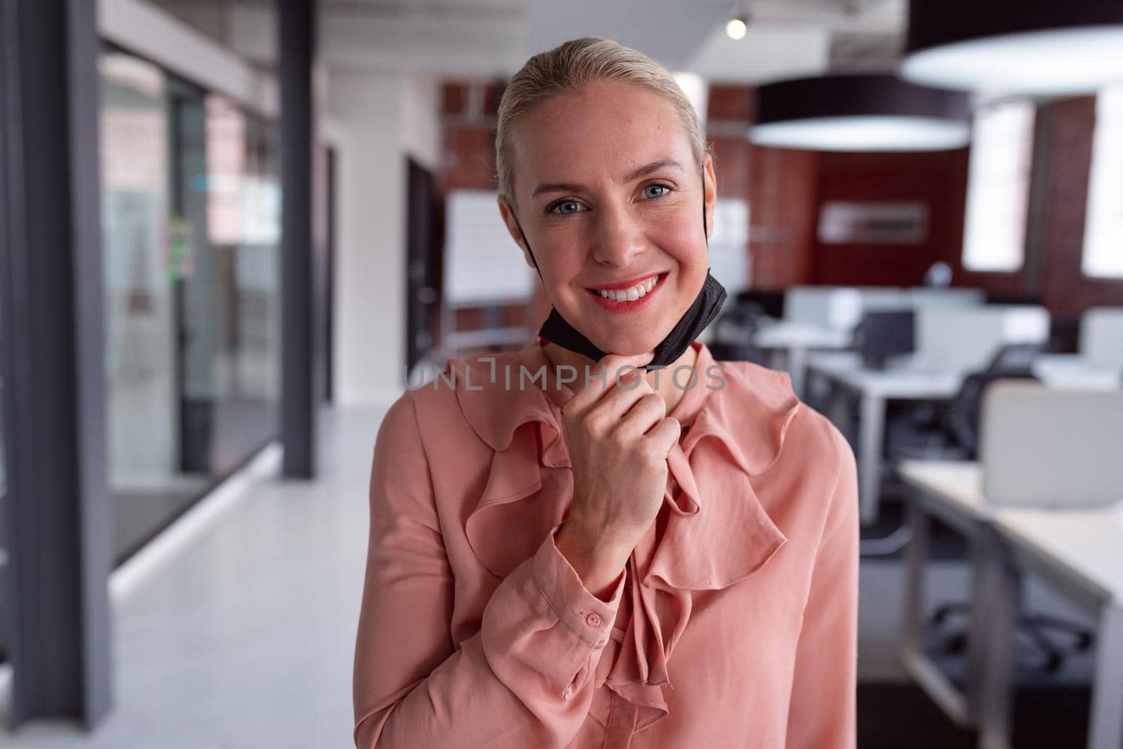 Portrait of caucasian businesswoman wearing lowered face mask standing in office smiling to camera. working in business at a modern office during coronavirus covid 19 pandemic.