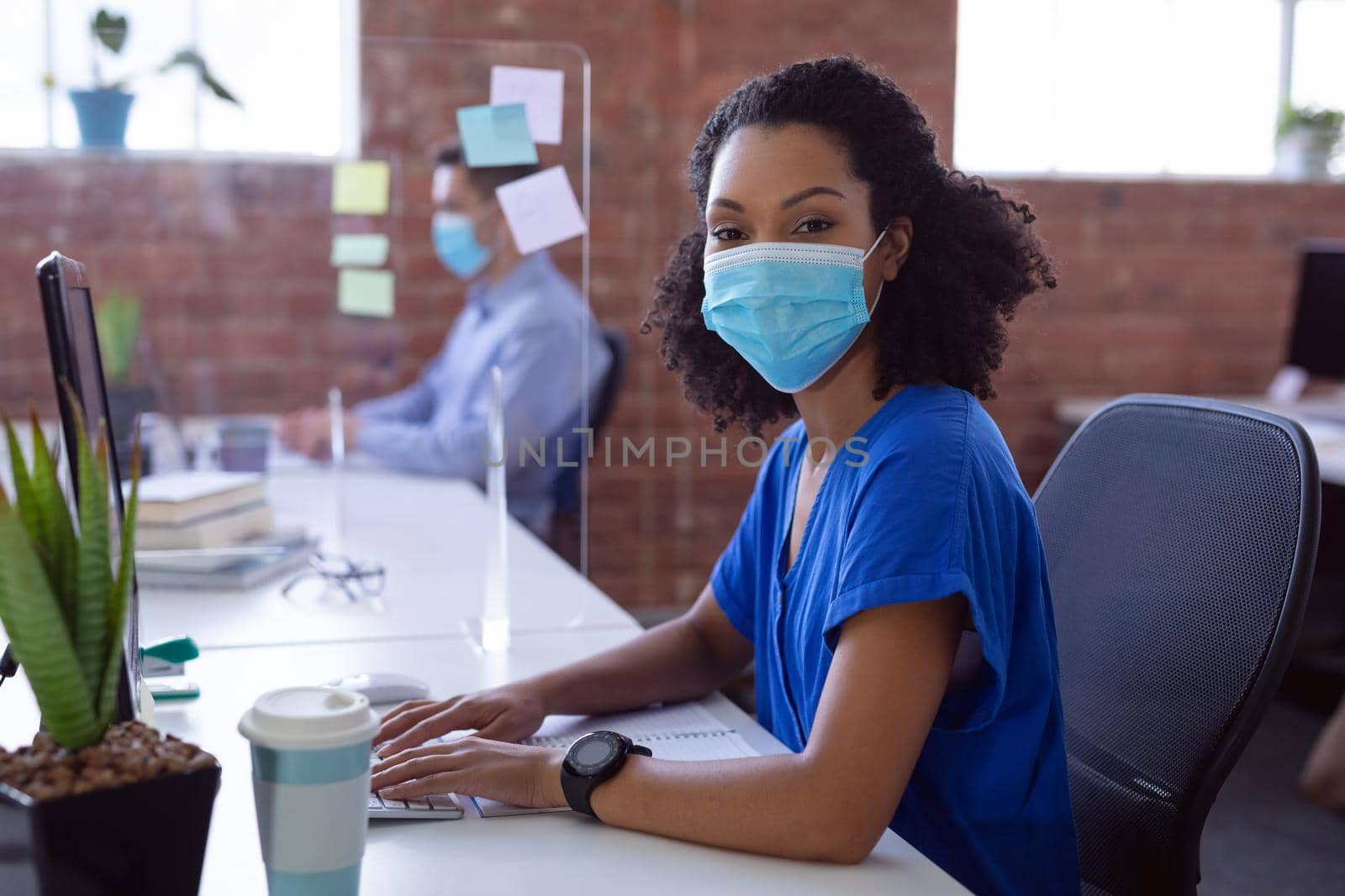 Portrait of african american businesswoman in face mask at desk with hygiene screen in office. working in business at a modern office during coronavirus covid 19 pandemic.