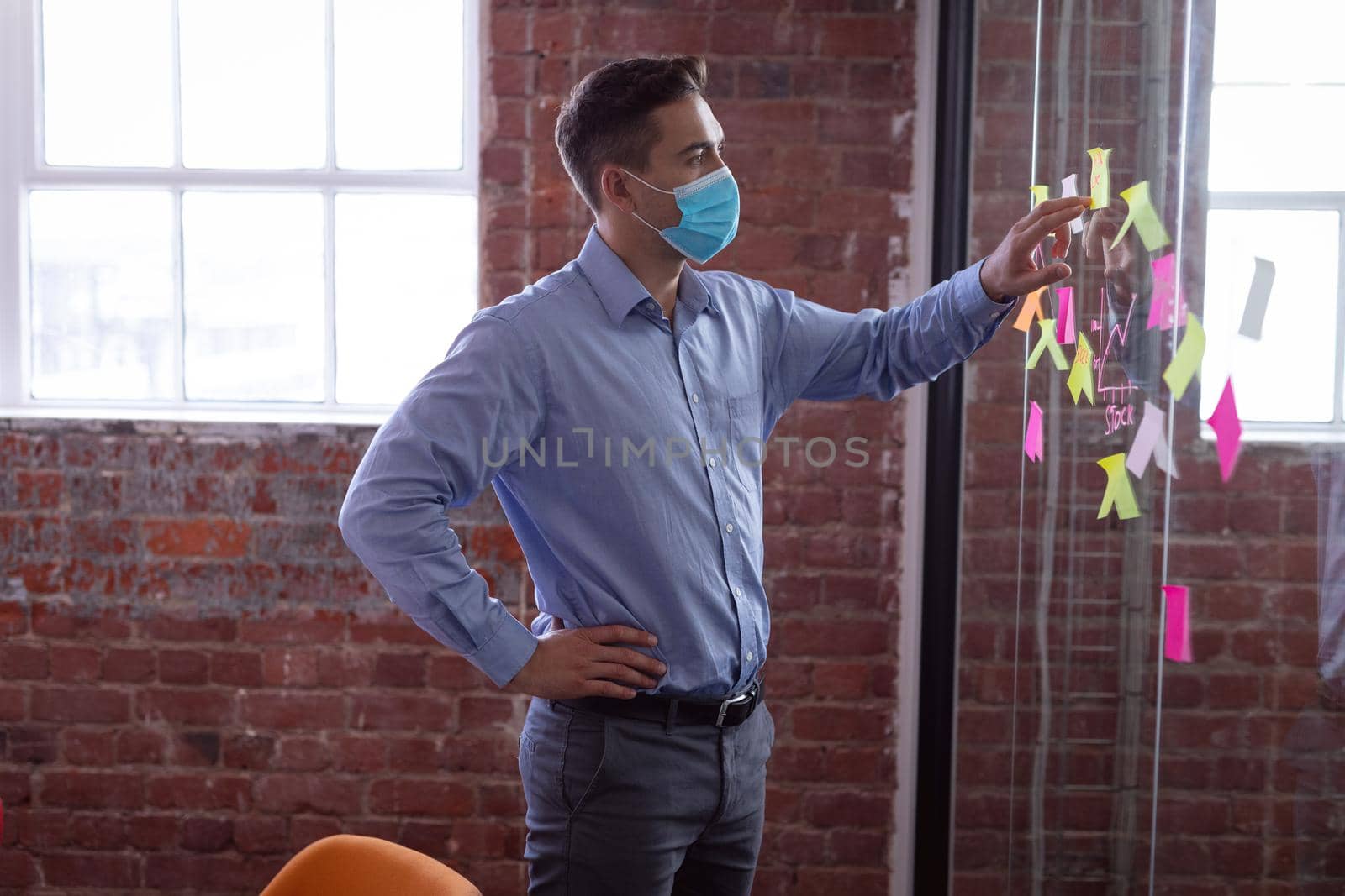 Caucasian businessman wearing face mask brainstorming, reading memo notes on wall standing in office by Wavebreakmedia