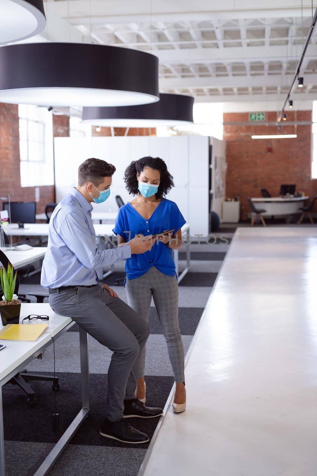 Diverse male and female colleague in face masks looking at tablet and discussing standing in office by Wavebreakmedia
