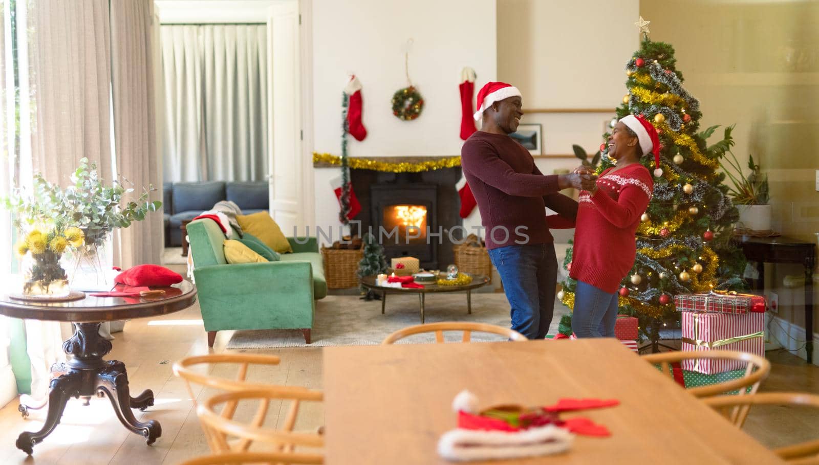 Happy african american senior couple in santa hats dancing at home at christmas time. retirement lifestyle and christmas festivities, celebrating at home.