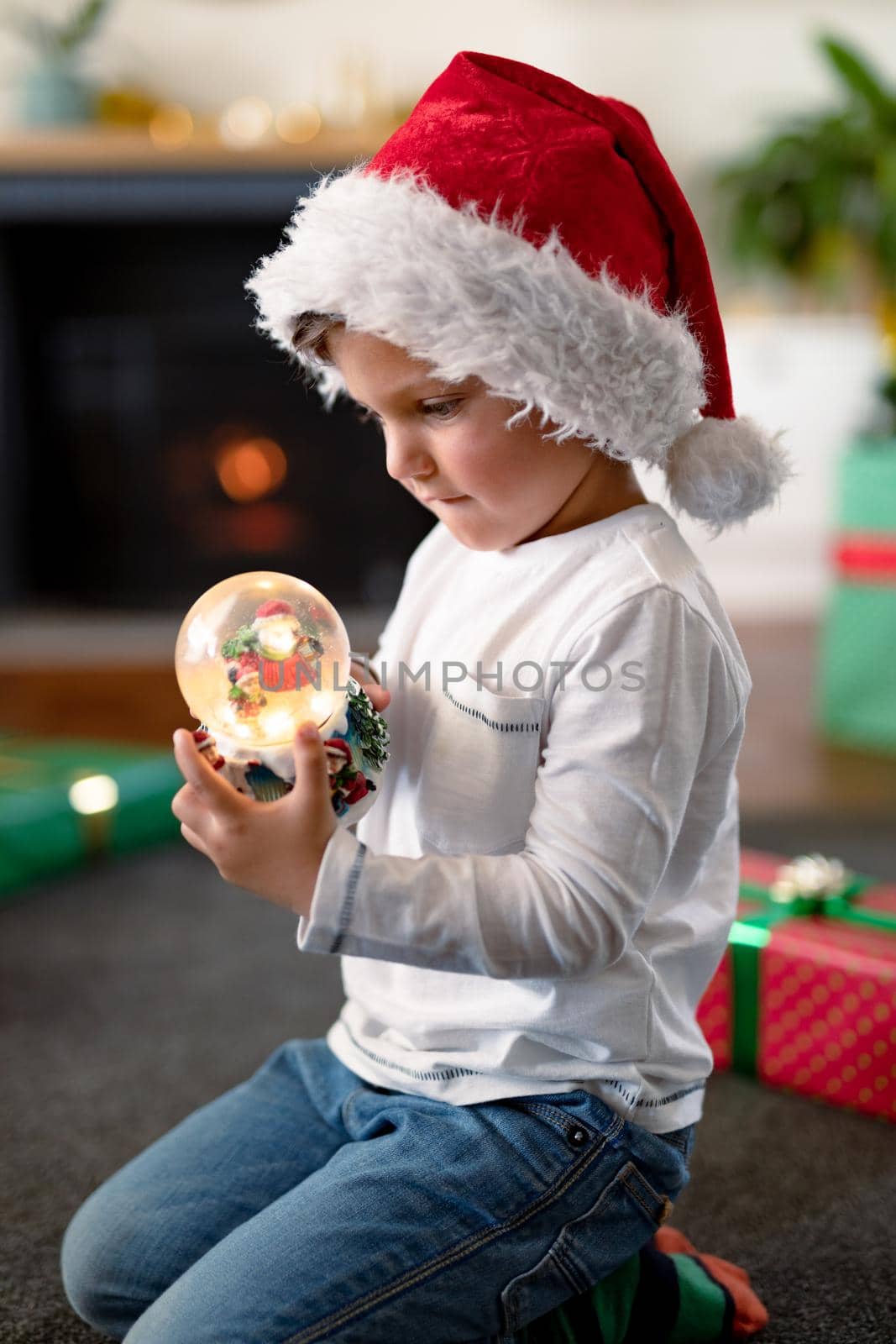 Focused caucasian boy wearing santa hat, looking at snow globe at christmas time. childhood, chiristmas, festivity and tradition at home.