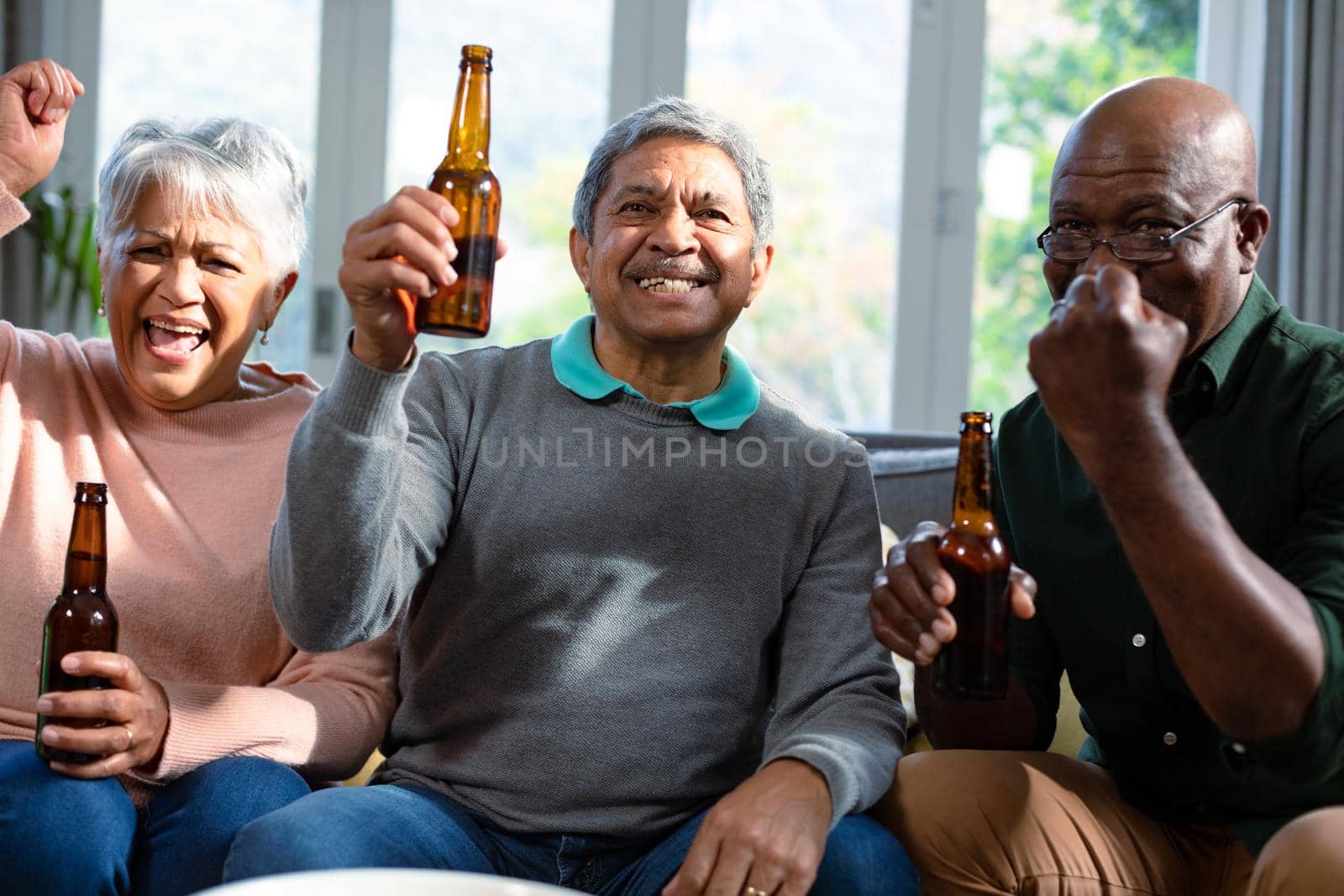 Two diverse senior couple and their african american friend watching tv and drinking beer. retirement lifestyle relaxing at home with technology.