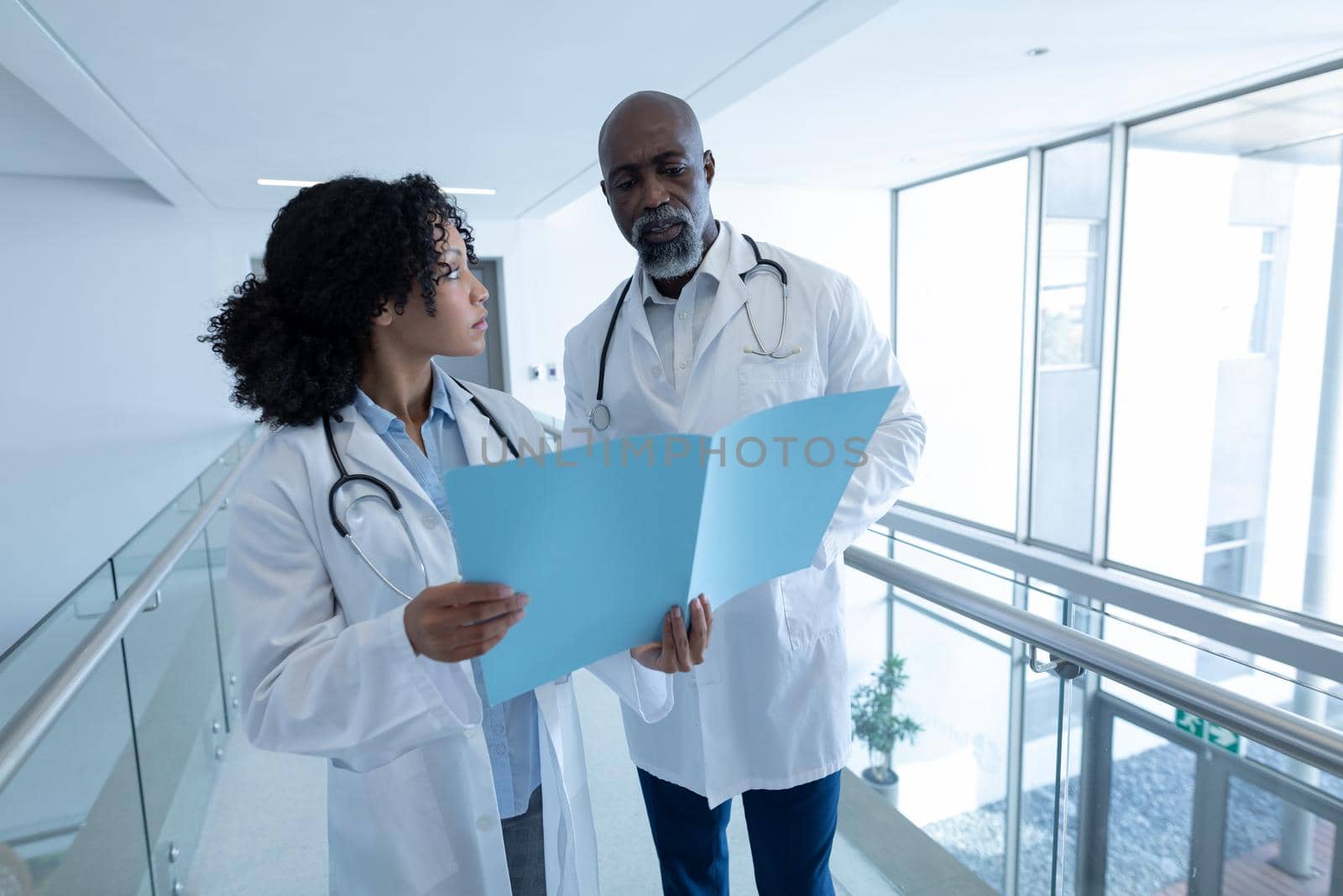 Diverse male and female doctor talking seriously and reading patient file in hospital corridor by Wavebreakmedia