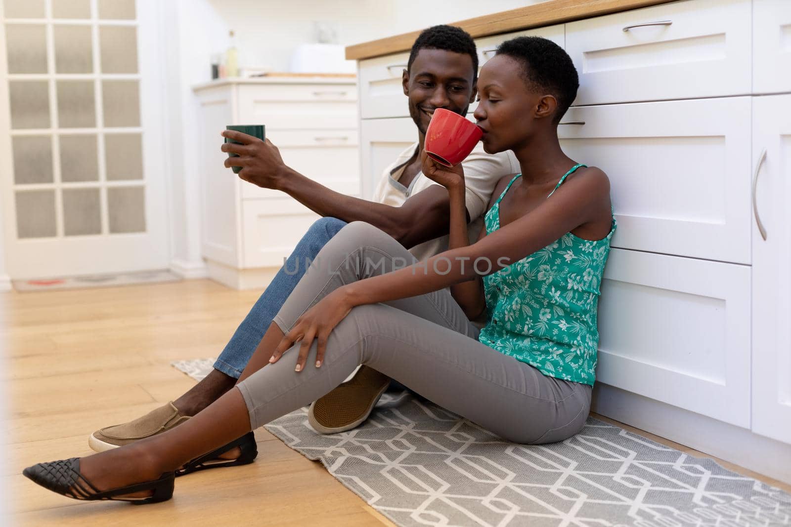 African american couple sitting on floor in kitchen holding mugs. staying at home in isolation during quarantine lockdown.