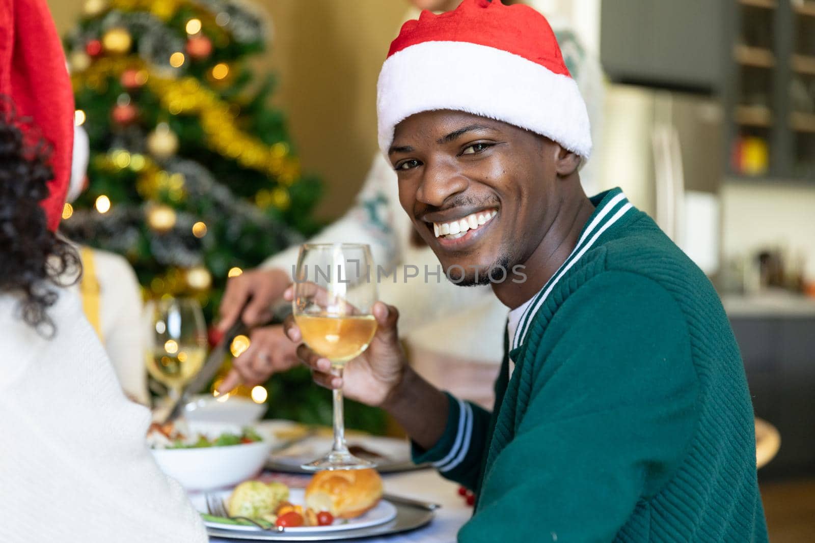 Happy african american man in santa hat toasting, celebrating christmas with friends at home. christmas festivities, celebrating at home with friends.