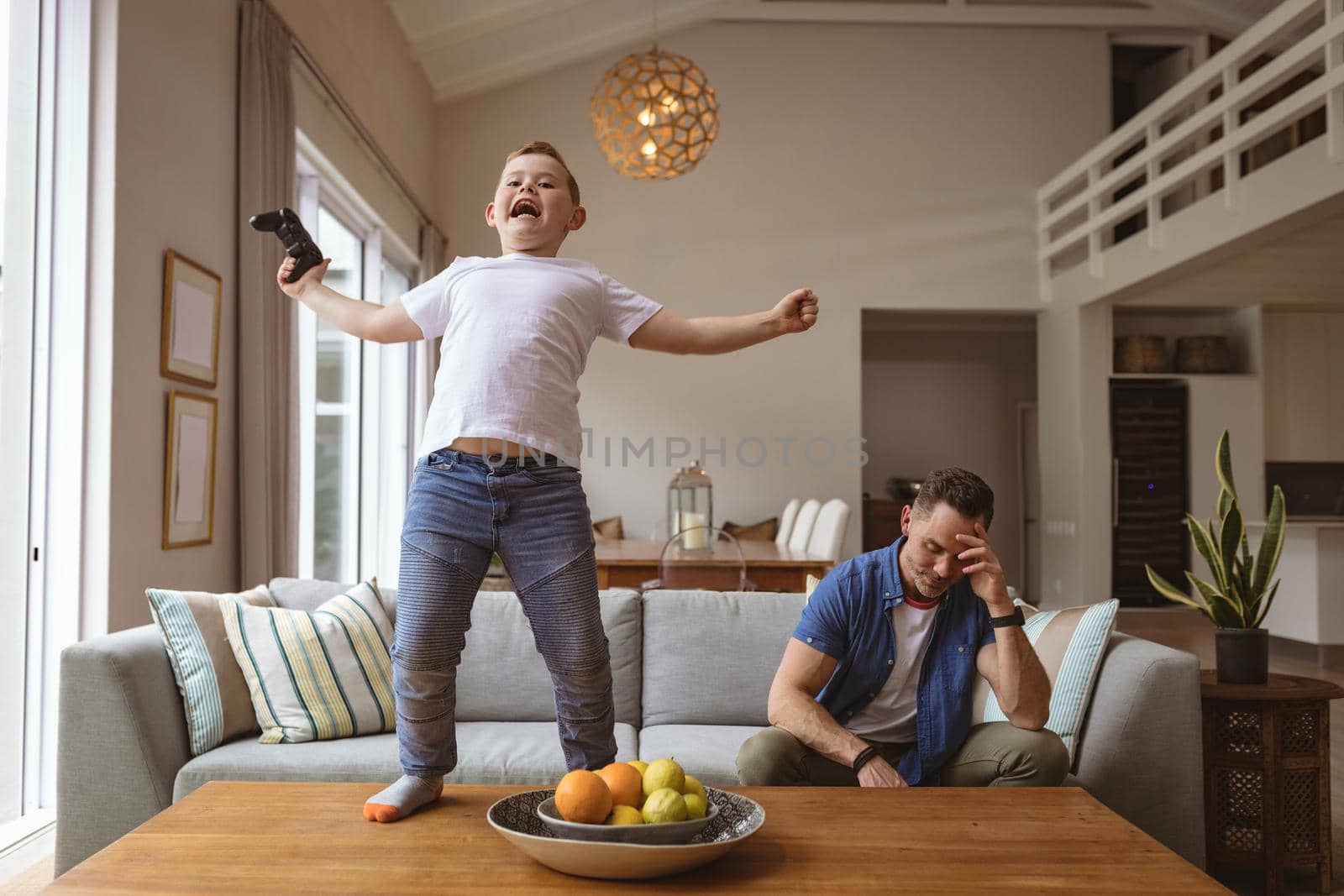 Caucasian boy celebrating standing on the table while playing video games with his father at home. gaming and entertainment concept