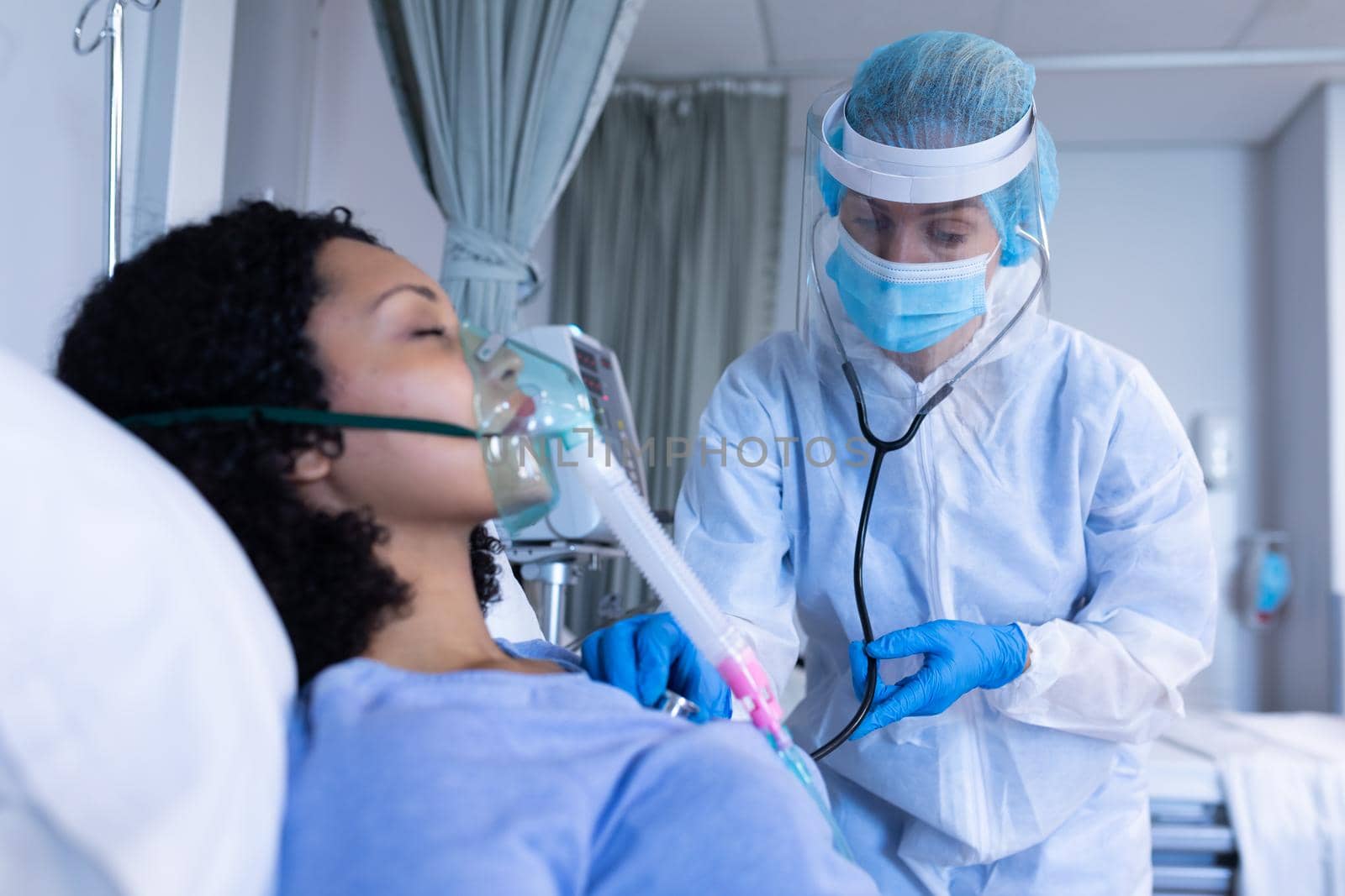 Caucasian doctor in ppe suit examining with stethoscope female patient with oxygen ventilator. medical professional at work during coronavirus covid 19 pandemic.