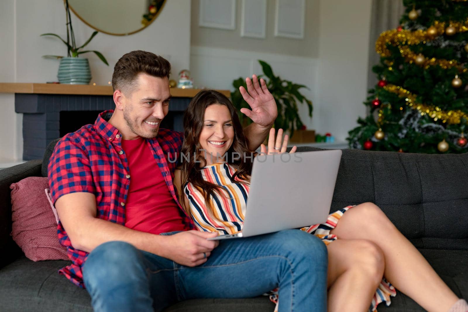 Happy caucasian couple sitting on sofa and having video call on laptop at christmas time. christmas, festivity and communication technology.
